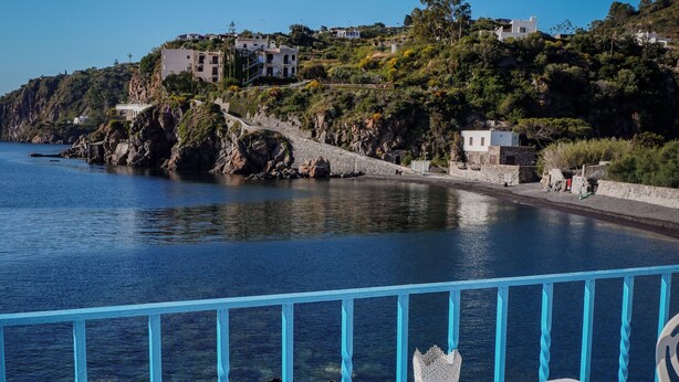 A table with a plate of food, a glass of red drink, and a fork, overlooking a coastal area with buildings and a clear blue sky. - Rocce Azzurre (Lipari)