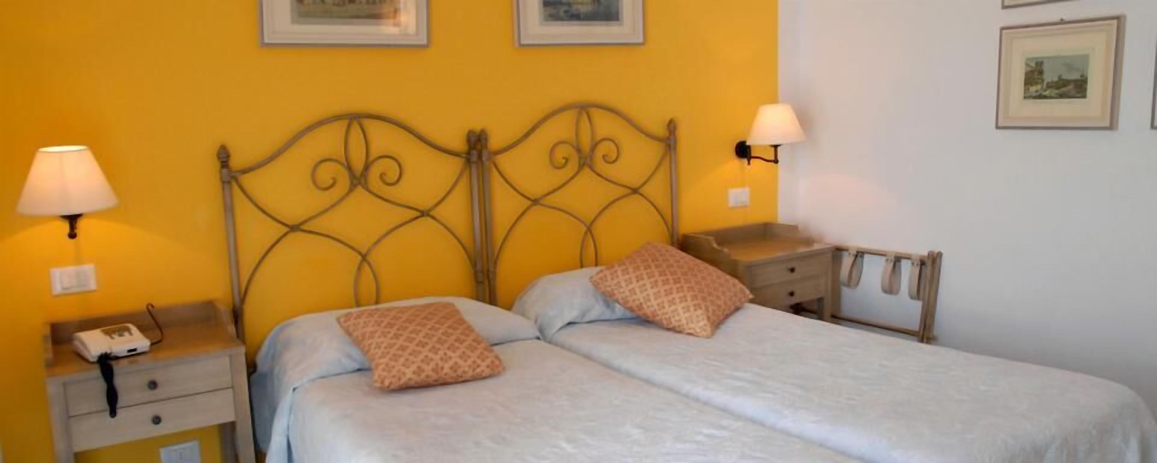 Una cama doble con cabecera de metal, dos mesitas de noche con lámparas, y una mesita auxiliar con lámpara.