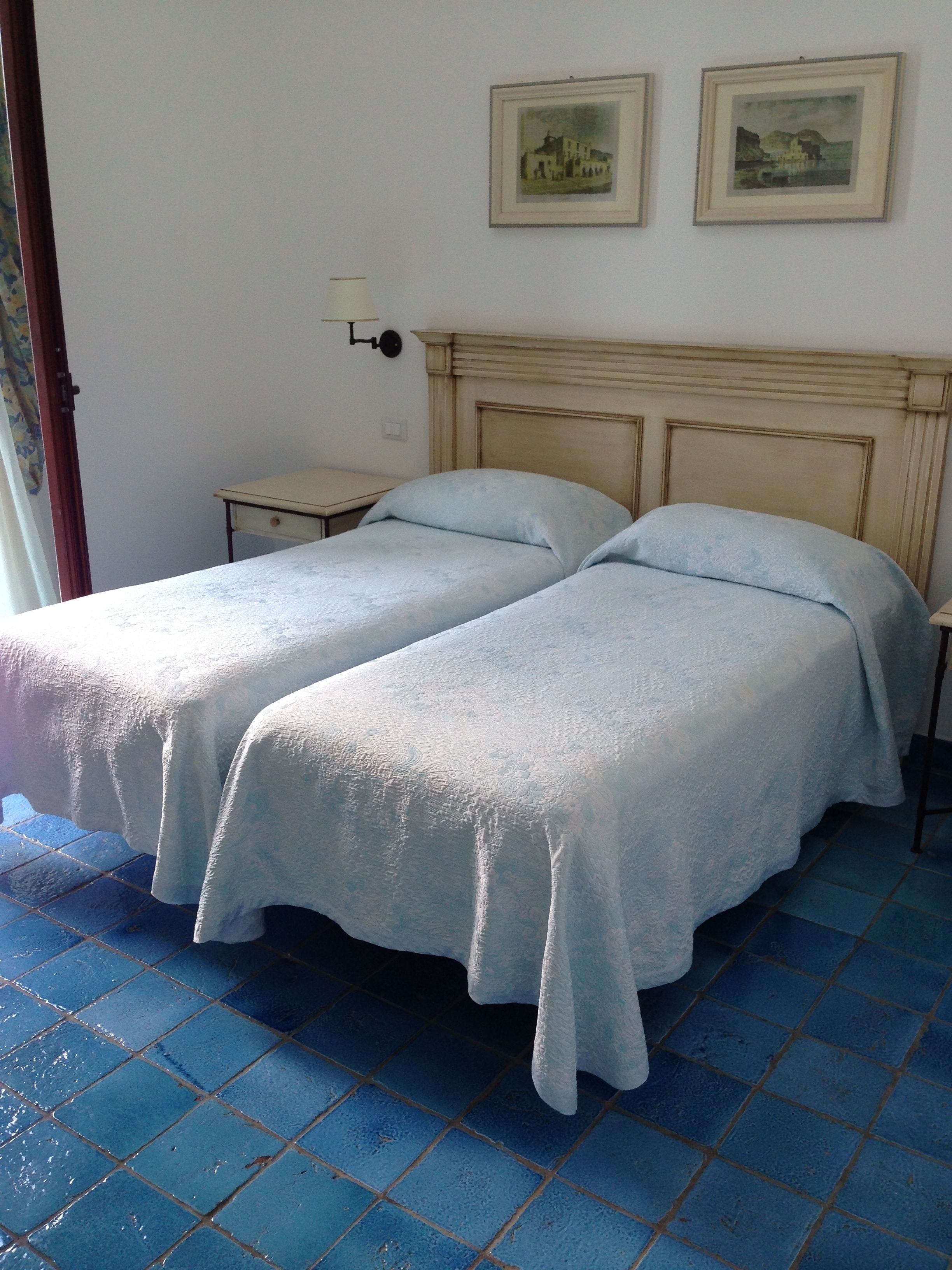 A room with two beds, a small bedside table, a wall-mounted lamp, and two framed pictures on the wall.