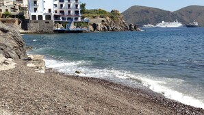 A pebbly beach with a white building with balconies, a cruise ship in the background, and hills in the distance. - Rocce Azzurre (Lipari)