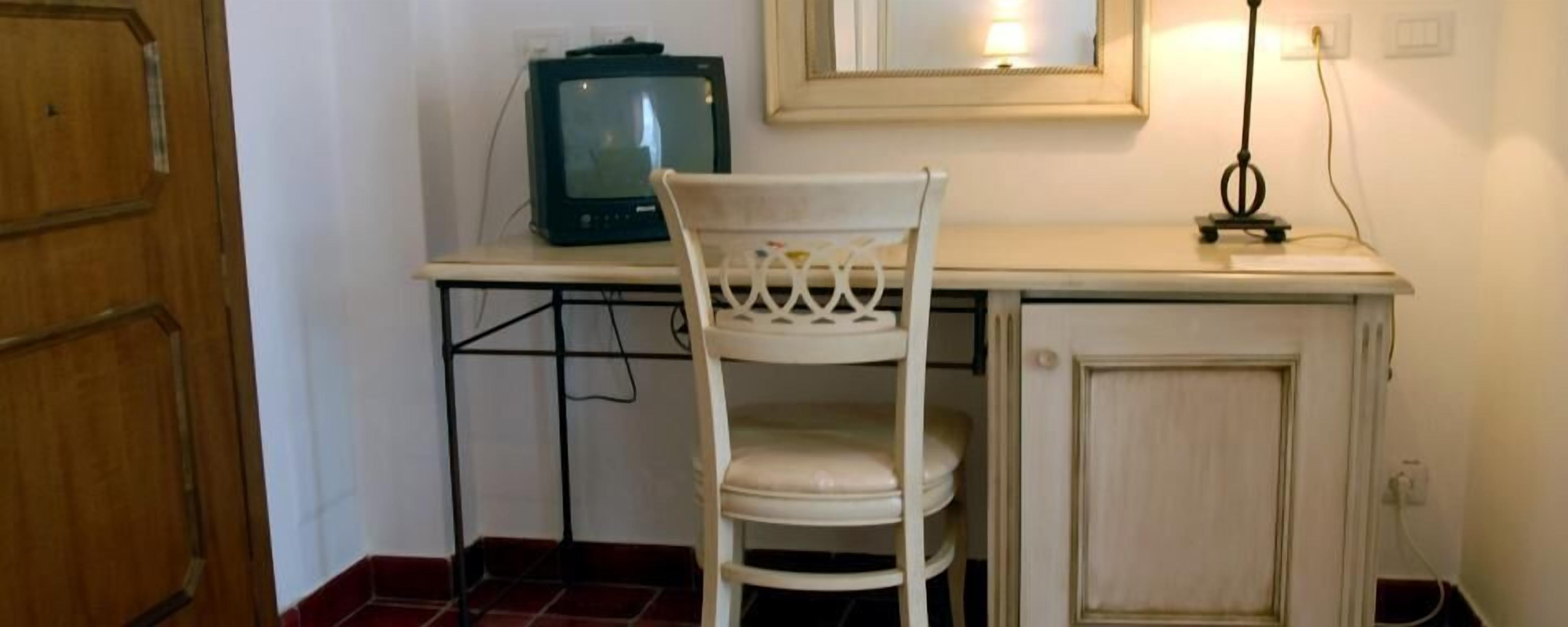 Una habitación pequeña con una puerta de madera, una silla blanca, un televisor sobre una mesa y un espejo en la pared.