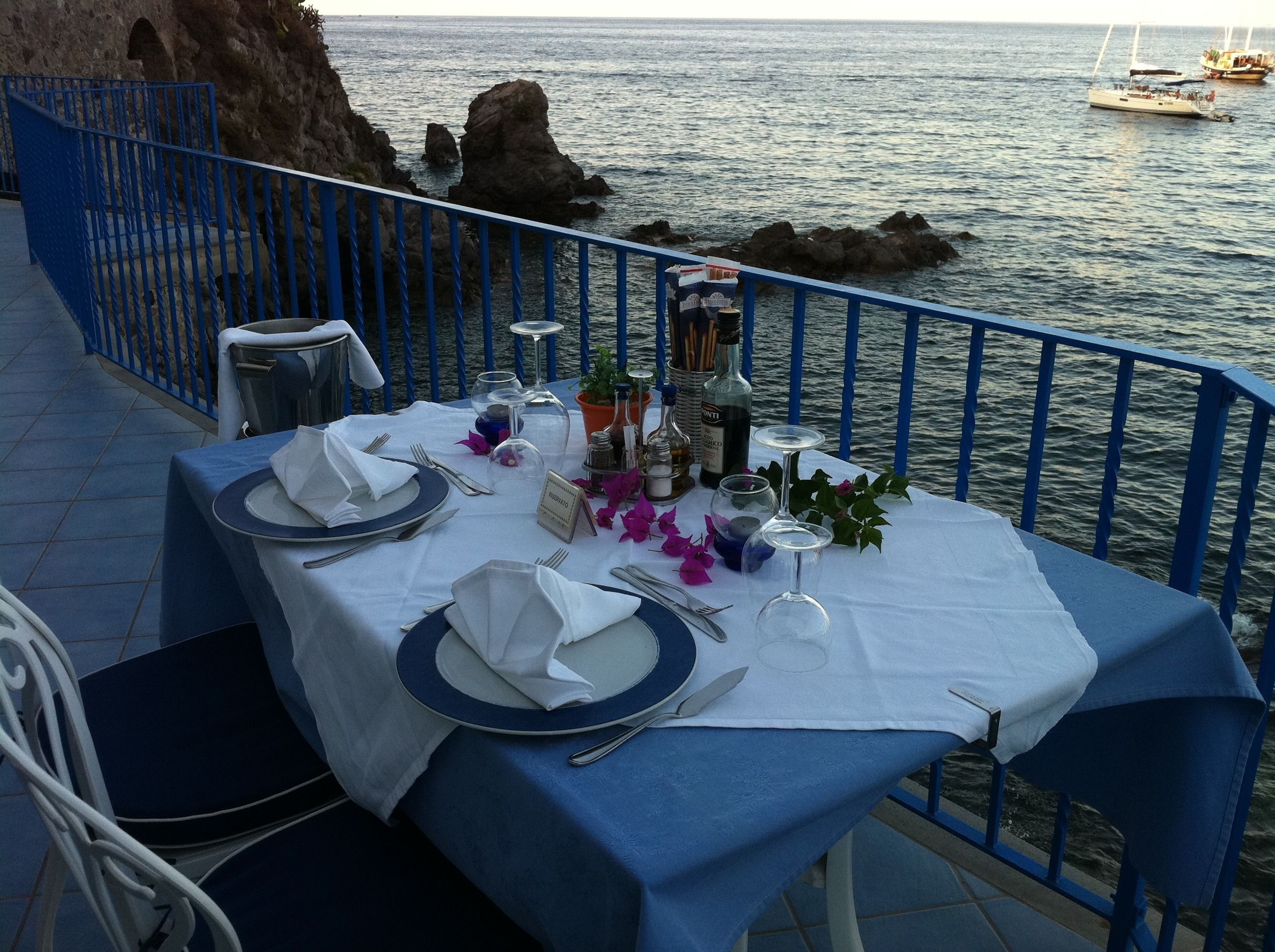 Tavolo imbandito per due con vista sul mare e sulla costa rocciosa.