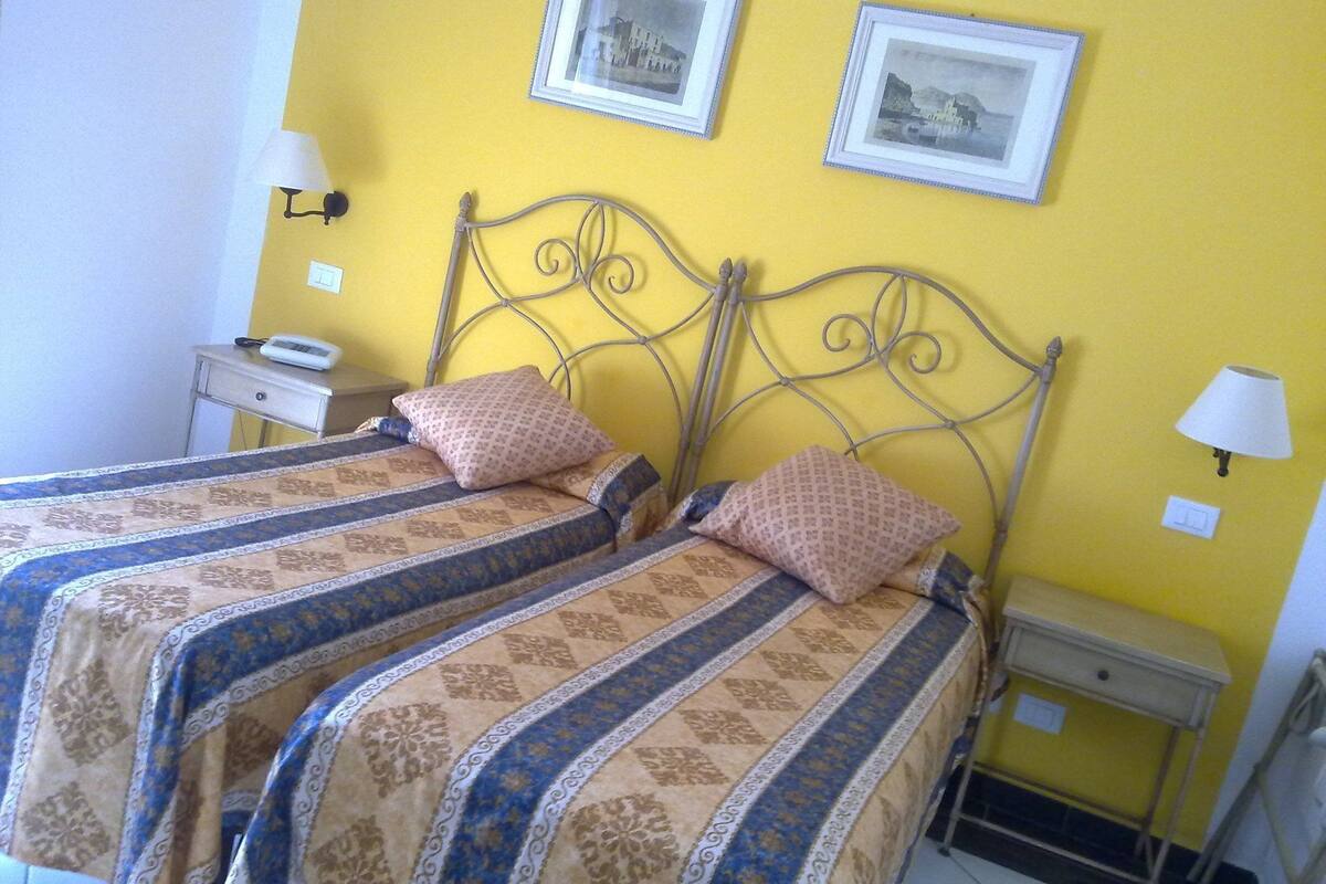 A double bed with patterned bedding, two wall-mounted lamps, and two framed pictures on the wall.
