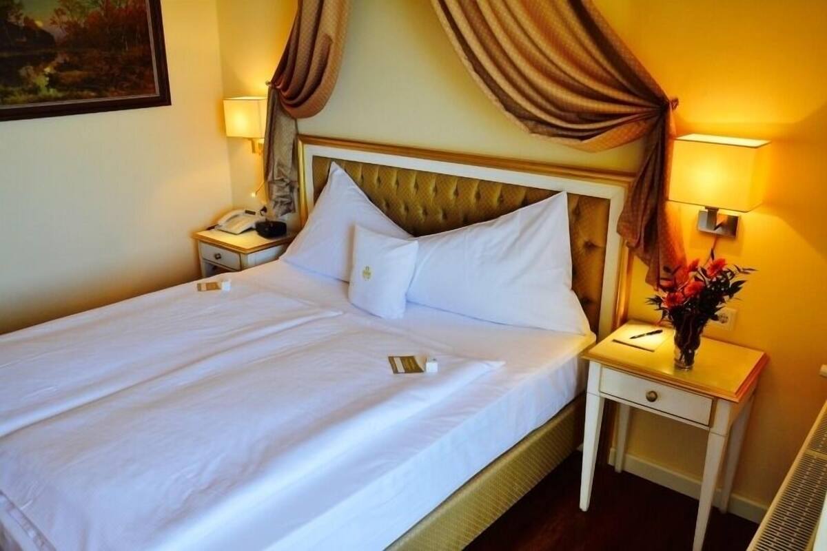 A hotel room with a bed, two bedside tables, a nightstand, a lamp, and a vase with flowers.