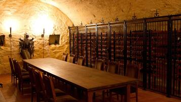 A stone-walled room with a long wooden table and chairs, metal railings, and a wine rack in the background.
