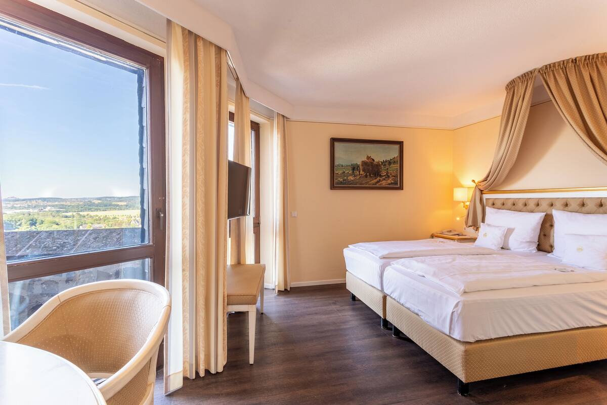 A hotel room with a large window, a bed, a chair, and a painting on the wall.