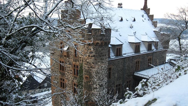 A historic stone building with multiple windows and a steeply pitched roof, surrounded by snow-covered trees and a hilly landscape.