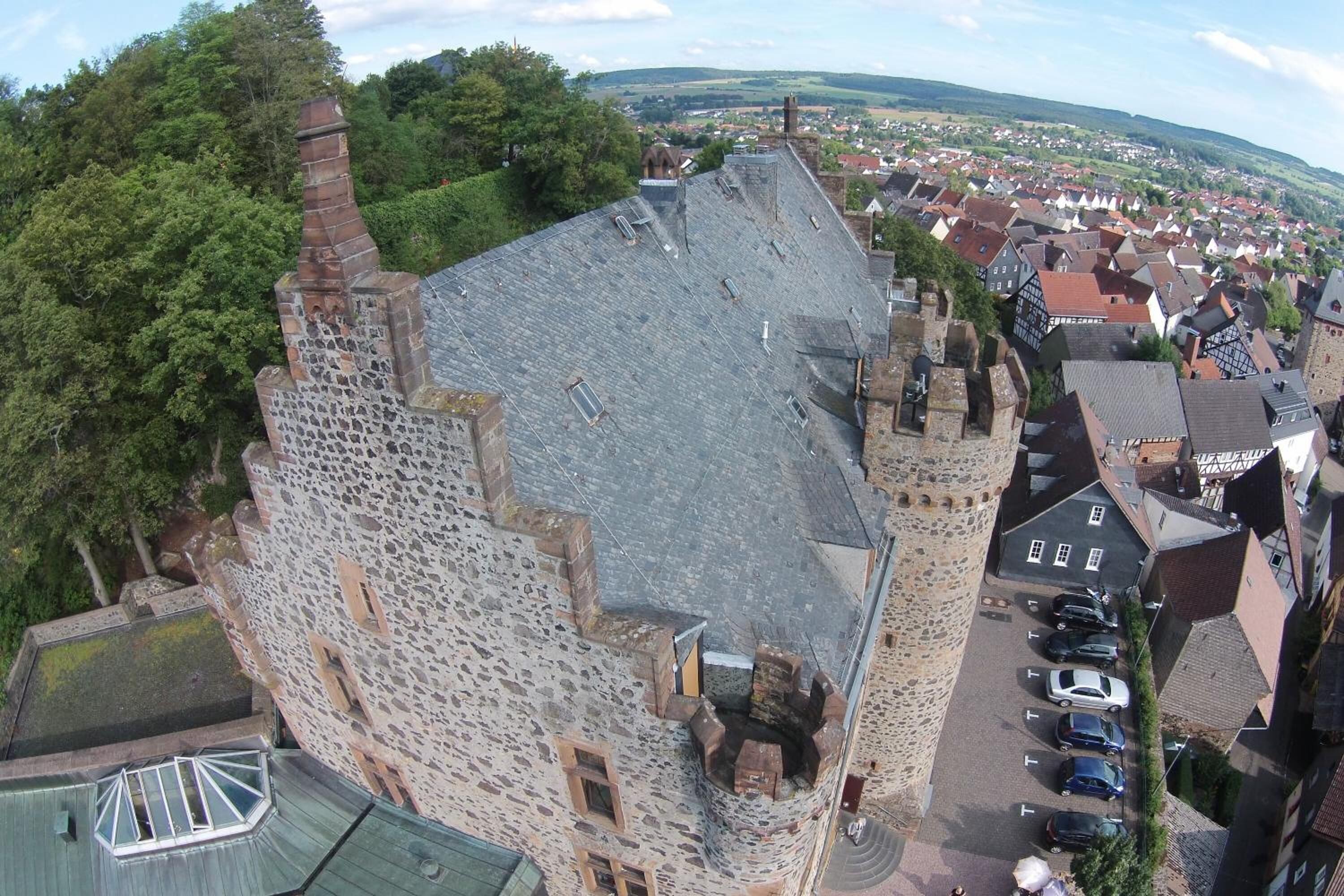 A historic stone building with a sloped roof, a courtyard with parked cars, and a view of a town in the background.