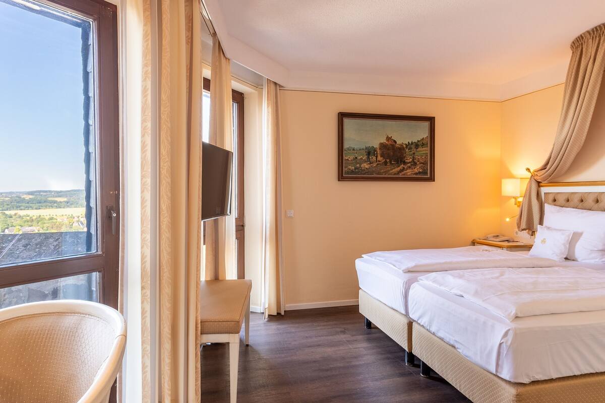 A hotel room with a bed, a chair, a painting, and a view of the countryside.