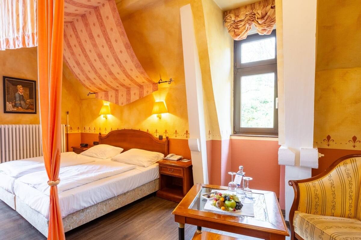 A hotel room with a four-poster bed, two bedside lamps, a wooden nightstand, a small table with fruit, and a chair.