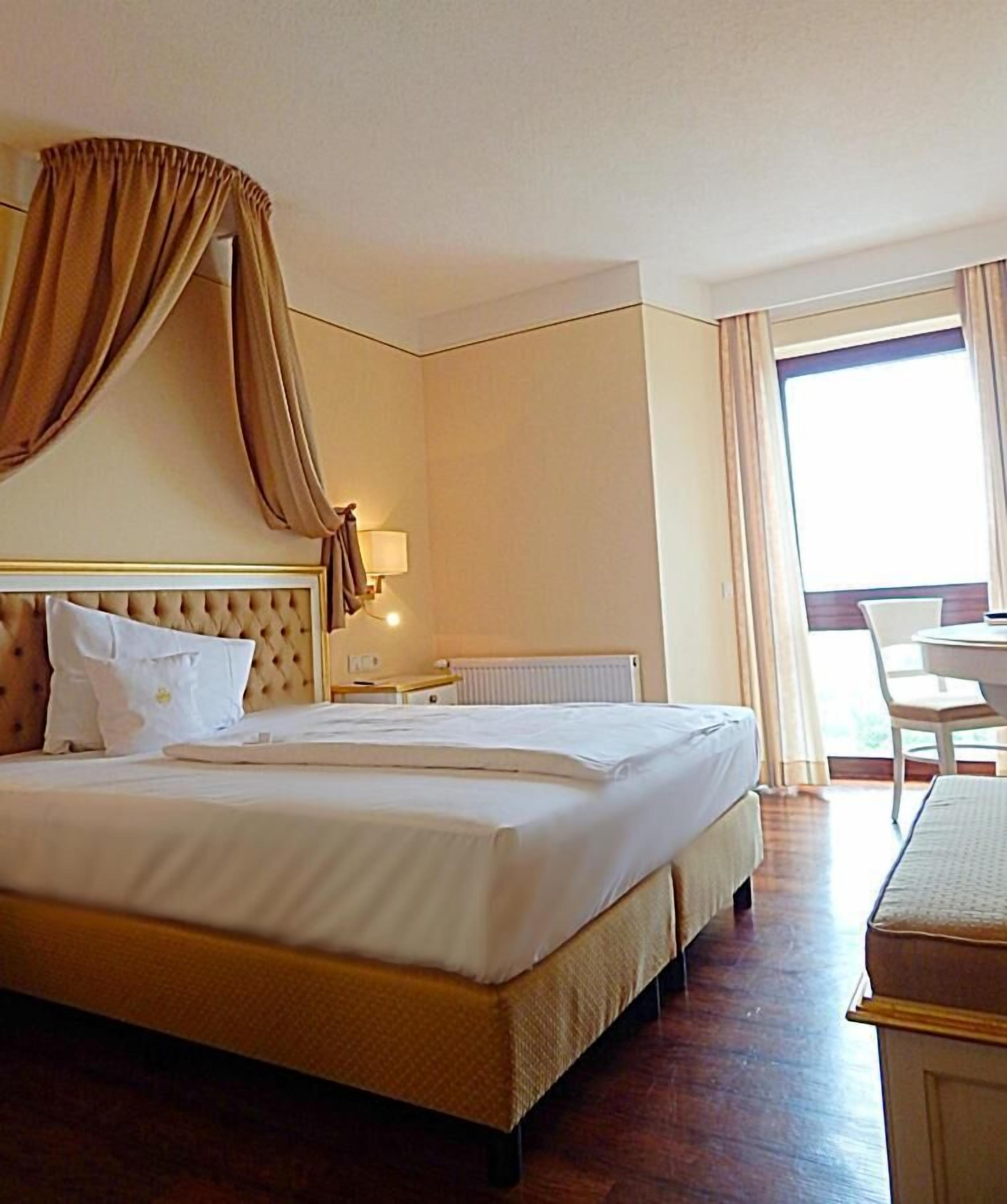 A hotel room with a large bed, a bedside table, a lamp, and a view of the outdoors.