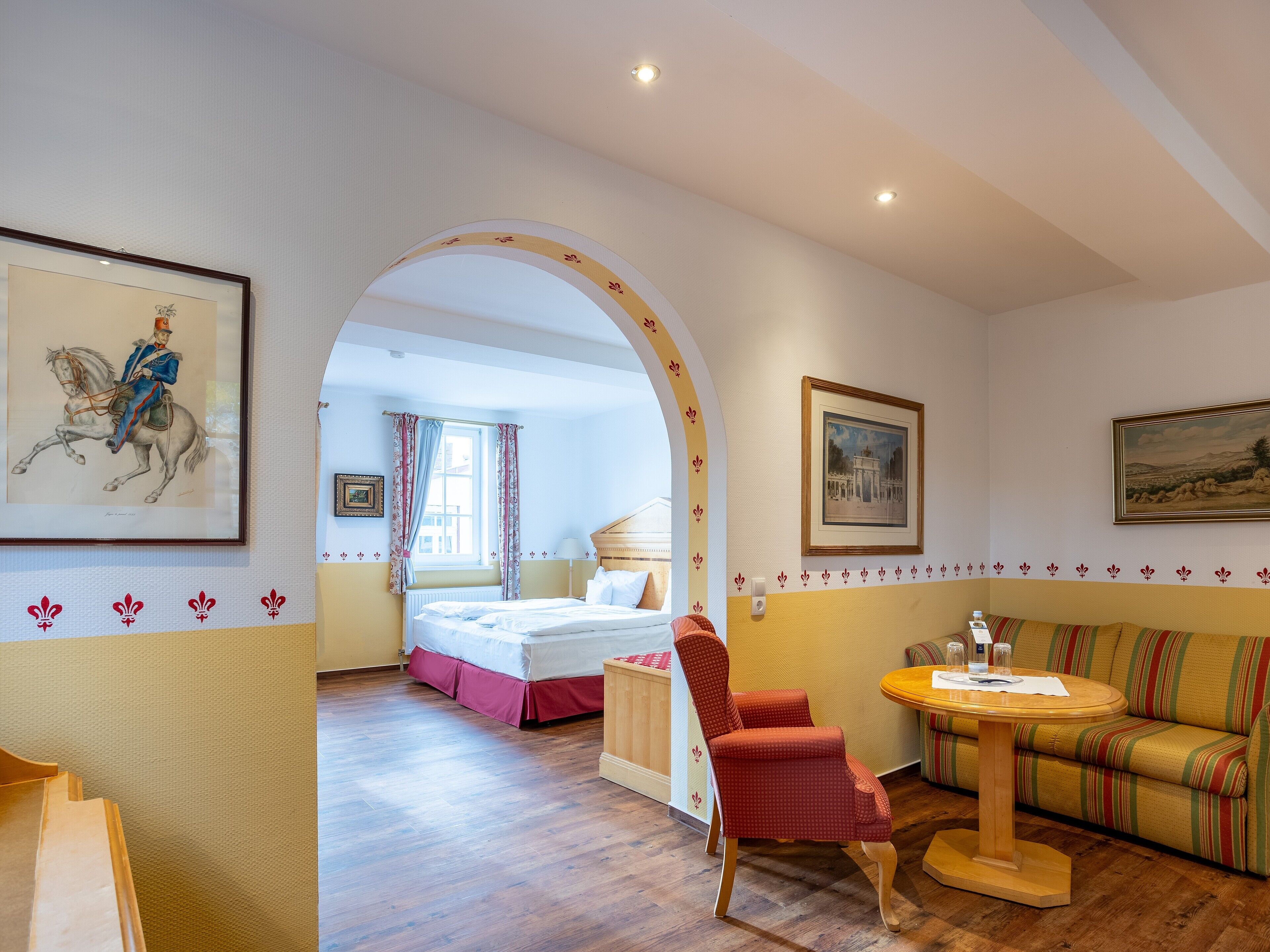 A hotel room with a bed, a red chair, a round table, and a sofa. There is a window with curtains and a painting on the wall.