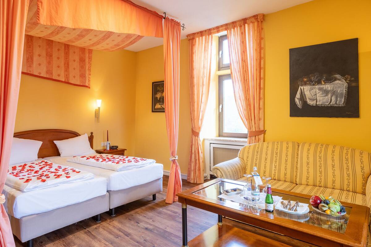 A hotel room with two beds, a sofa, a dining table with fruit and a bottle, and a painting on the wall.