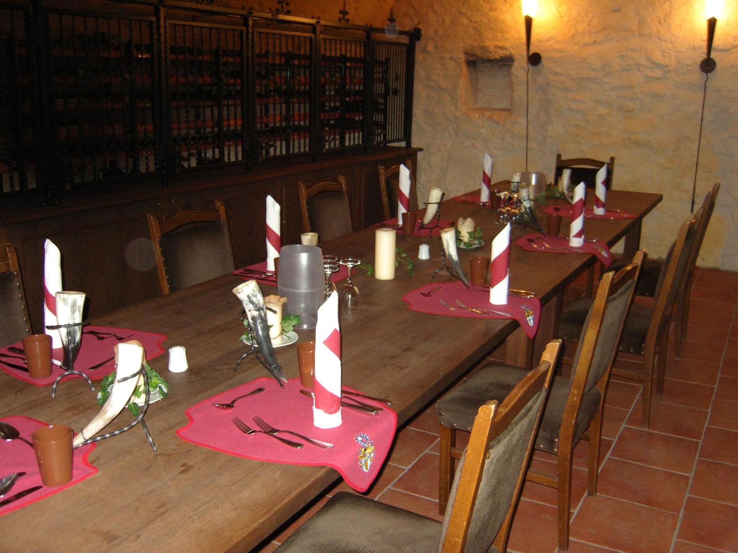 A long dining table set with red placemats, candles, and cutlery, surrounded by wooden chairs in a room with stone walls and a bookshelf in the background.