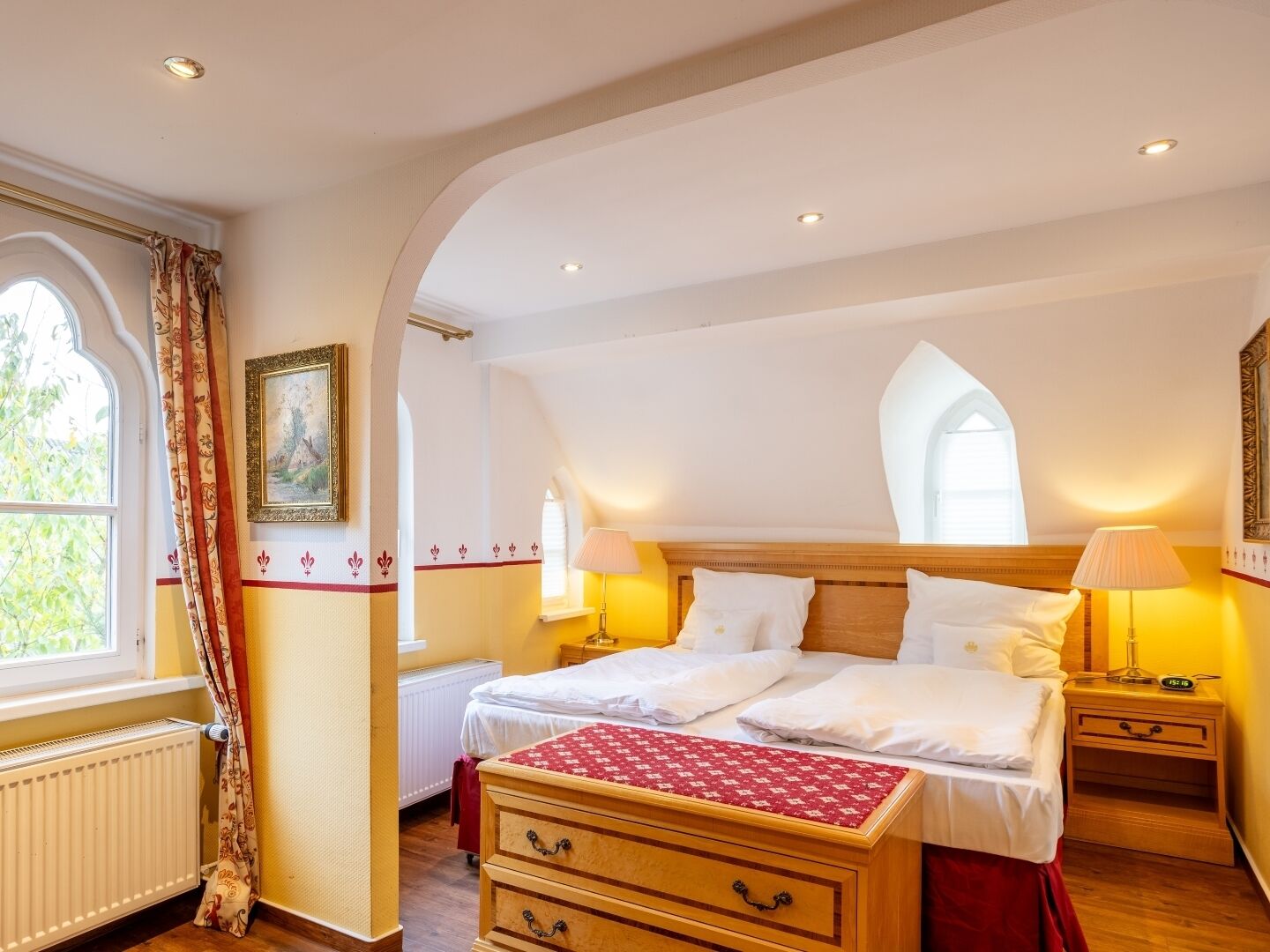 A bedroom with a double bed, two bedside tables with lamps, a wooden dresser, a radiator, and a window with curtains.