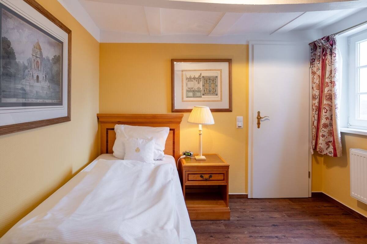 A neatly made bed with white linens, a wooden nightstand with a lamp, a framed picture on the wall, and a door with patterned curtains.