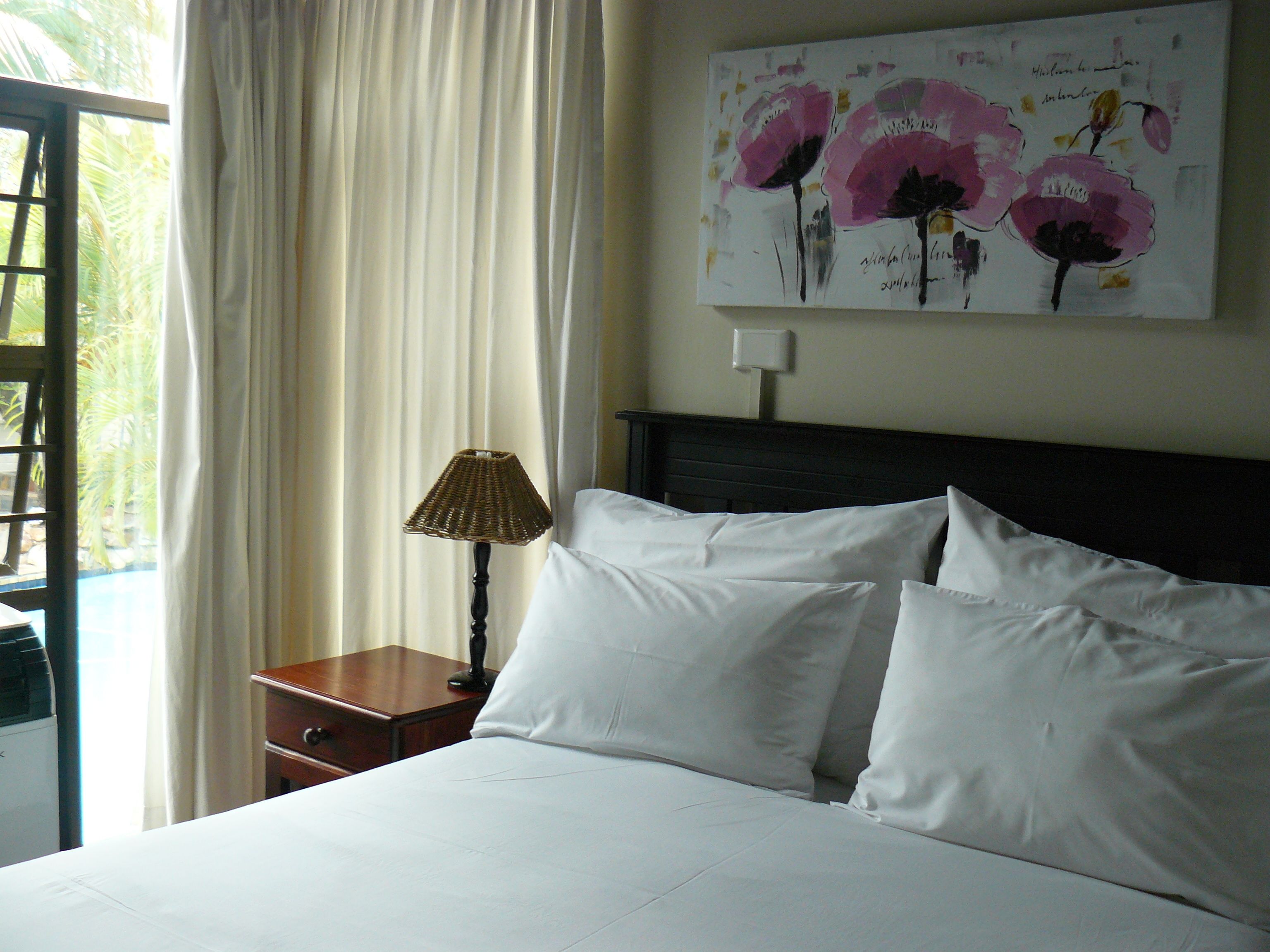 A hotel room with a bed, white pillows, a nightstand with a lamp, and a painting of flowers on the wall.