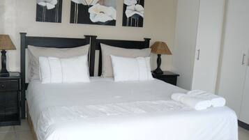 Uma cama bem arrumada com lençóis brancos, duas mesas de cabeceira com abajures e três quadros pendurados na parede retratando flores brancas.