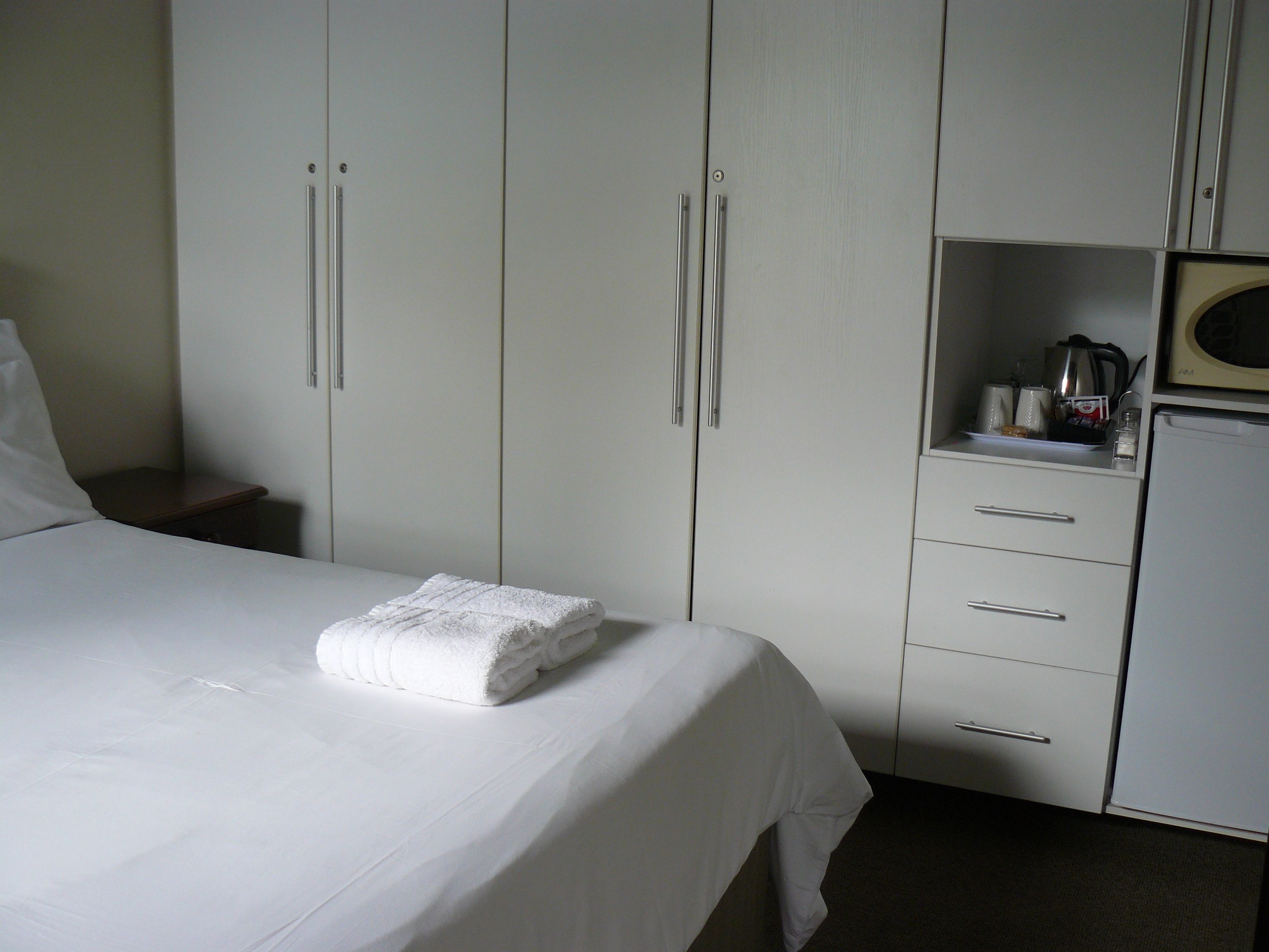 A hotel room with a neatly made bed, a small bedside table, a wardrobe with multiple doors, and a kitchenette area with a microwave.