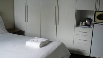 A hotel room with a neatly made bed, a small bedside table, a wardrobe with multiple doors, and a kitchenette area with a microwave.