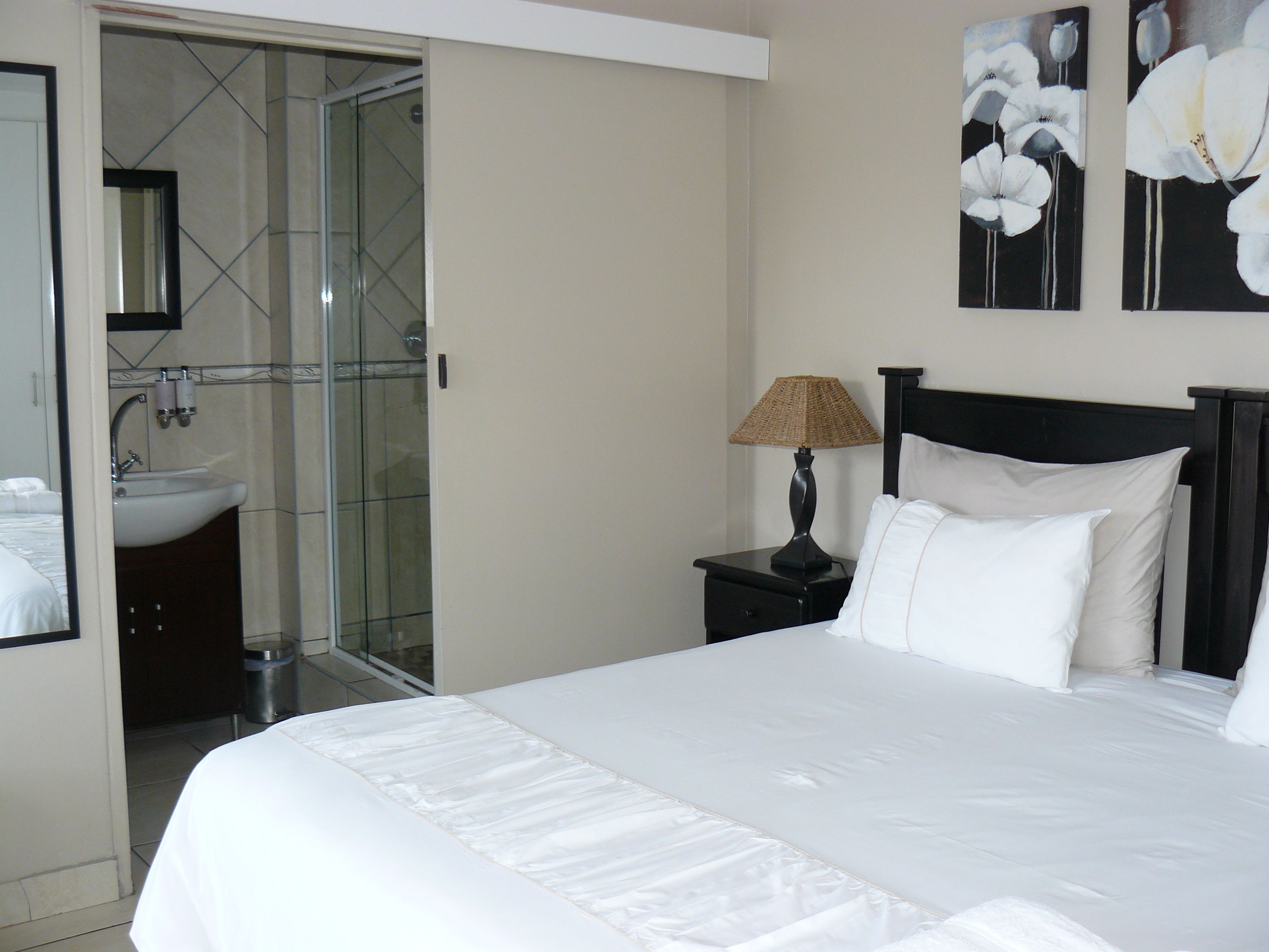 A hotel room with a large bed, a bedside table with a lamp, a bathroom with a glass shower enclosure, and two paintings on the wall.