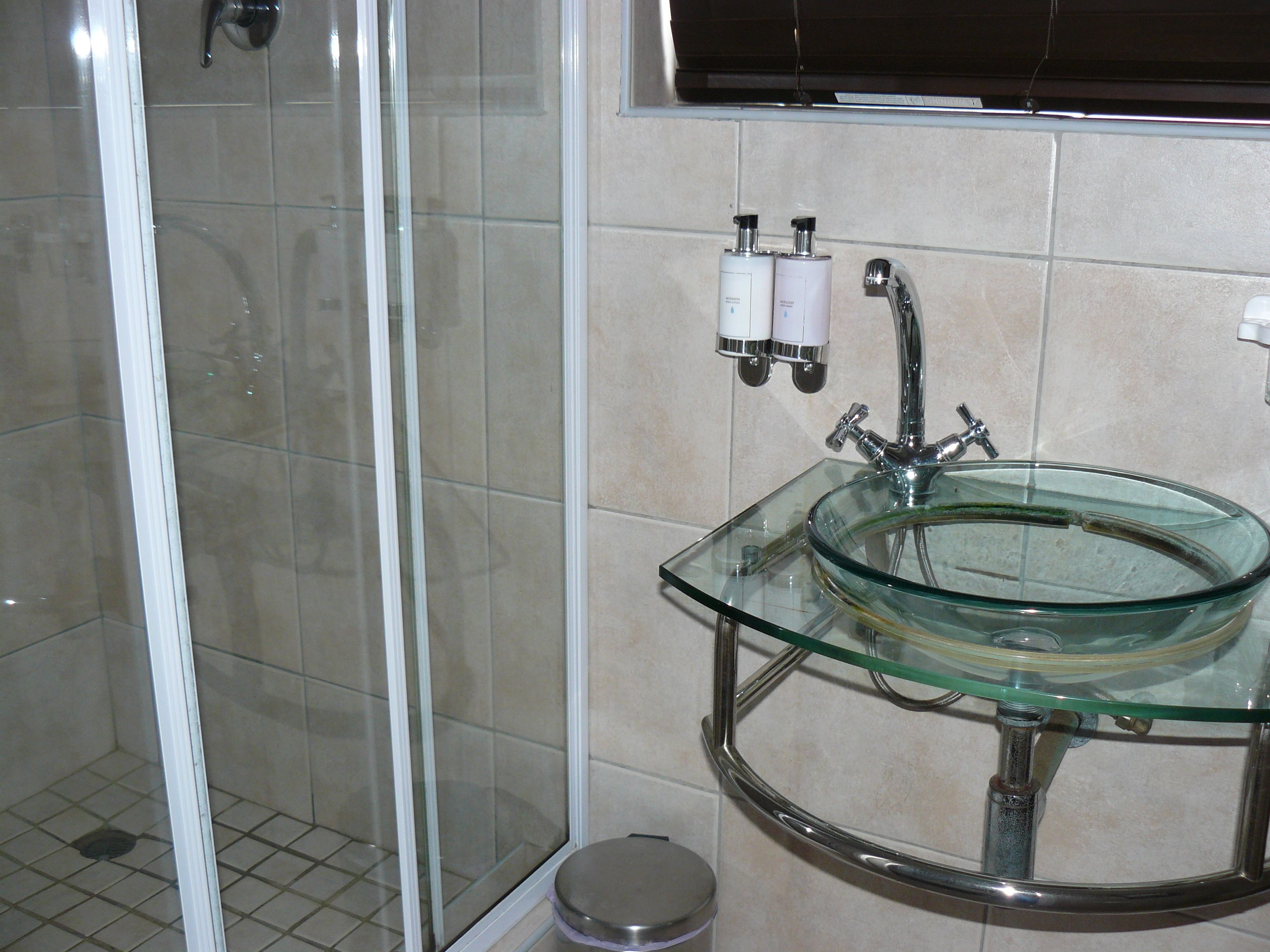A bathroom with a glass sink, a shower, and two bottles of shampoo on the wall.