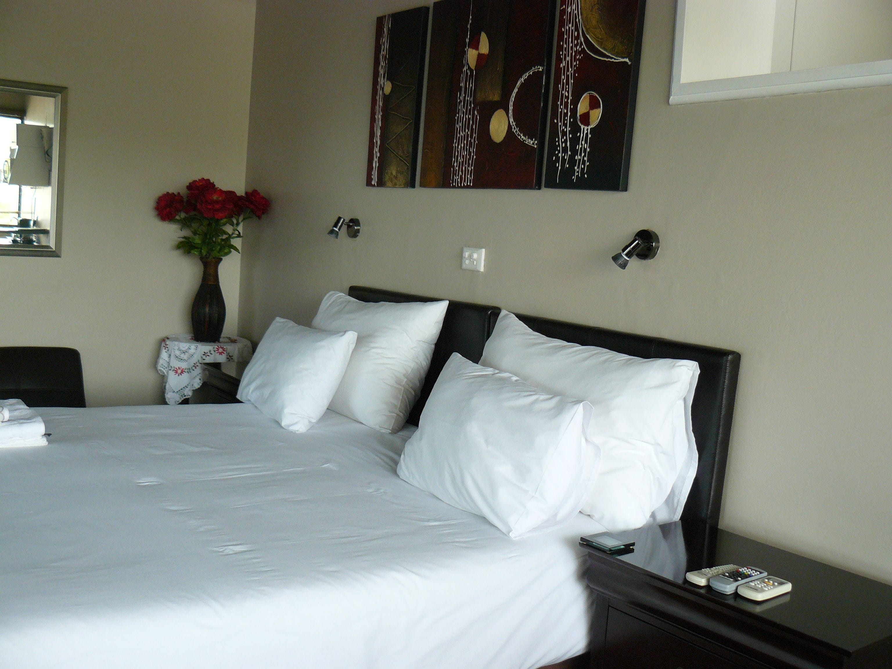 A neatly made bed with white linens, a vase with red flowers, and a painting on the wall.