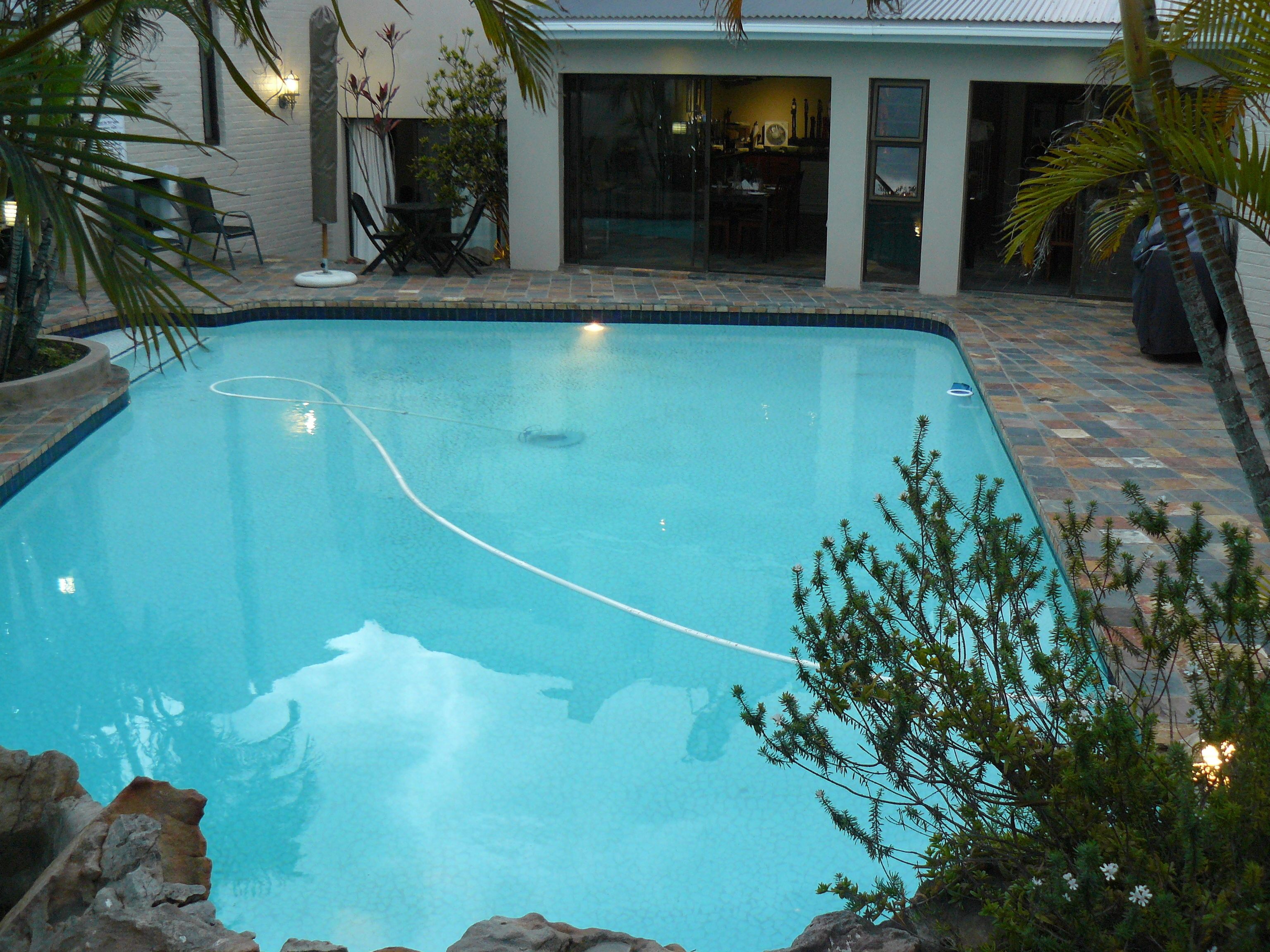 A rectangular swimming pool with a rope in the center, surrounded by a patio area with chairs and tables, and a building with large windows in the background.
