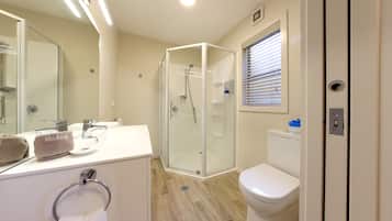 Un baño moderno con ducha de vidrio, lavamanos blanco y un toallón de maquillaje, y un inodoro con una ventana arriba.