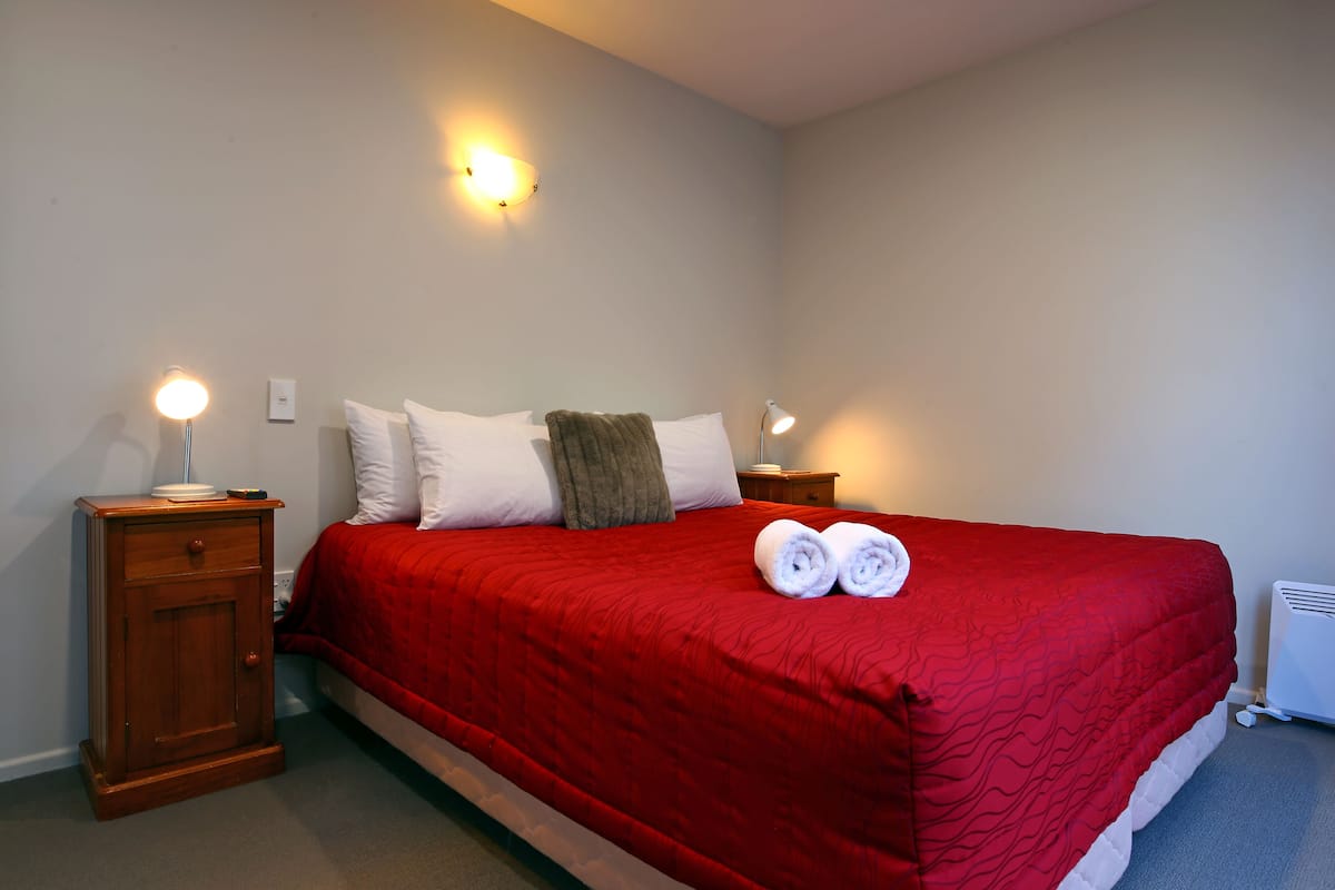 Ein ordentlich bezogenes Bett mit roter Tagesdecke, zwei weißen Kissen und einer braunen Decke. Auf den hölzernen Nachttischen stehen jeweils zwei Nachttischlampen.