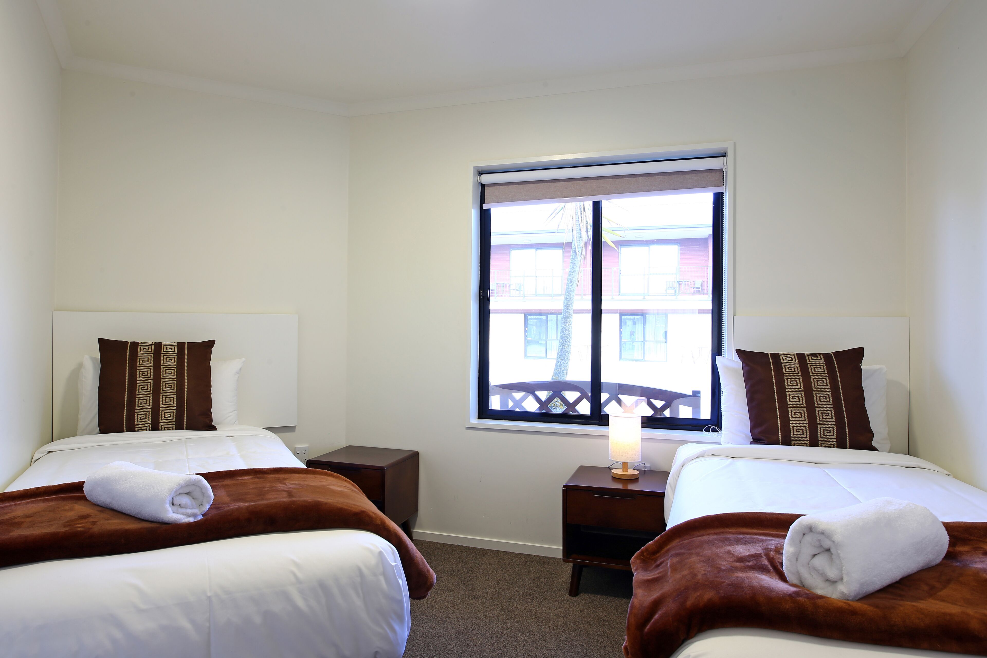 A hotel room with two beds, each with a brown headboard, white bedding, and a rolled towel on the bed. There is a window with blinds, a nightstand with a lamp, and a view of buildings outside.