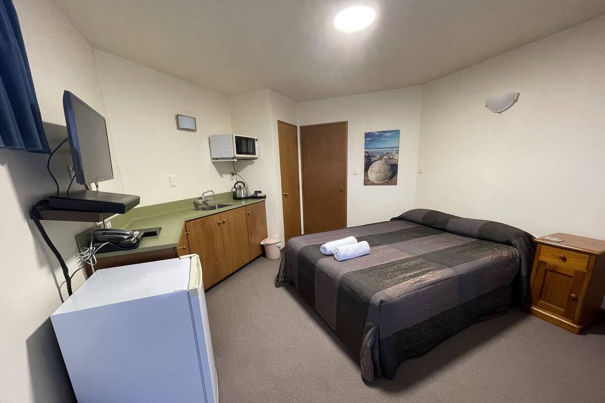 Ein kompaktes Hotelzimmer mit einem Einzelbett, einem kleinen Schreibtisch mit Computer, einer Küchenzeile und einem Badezimmer.