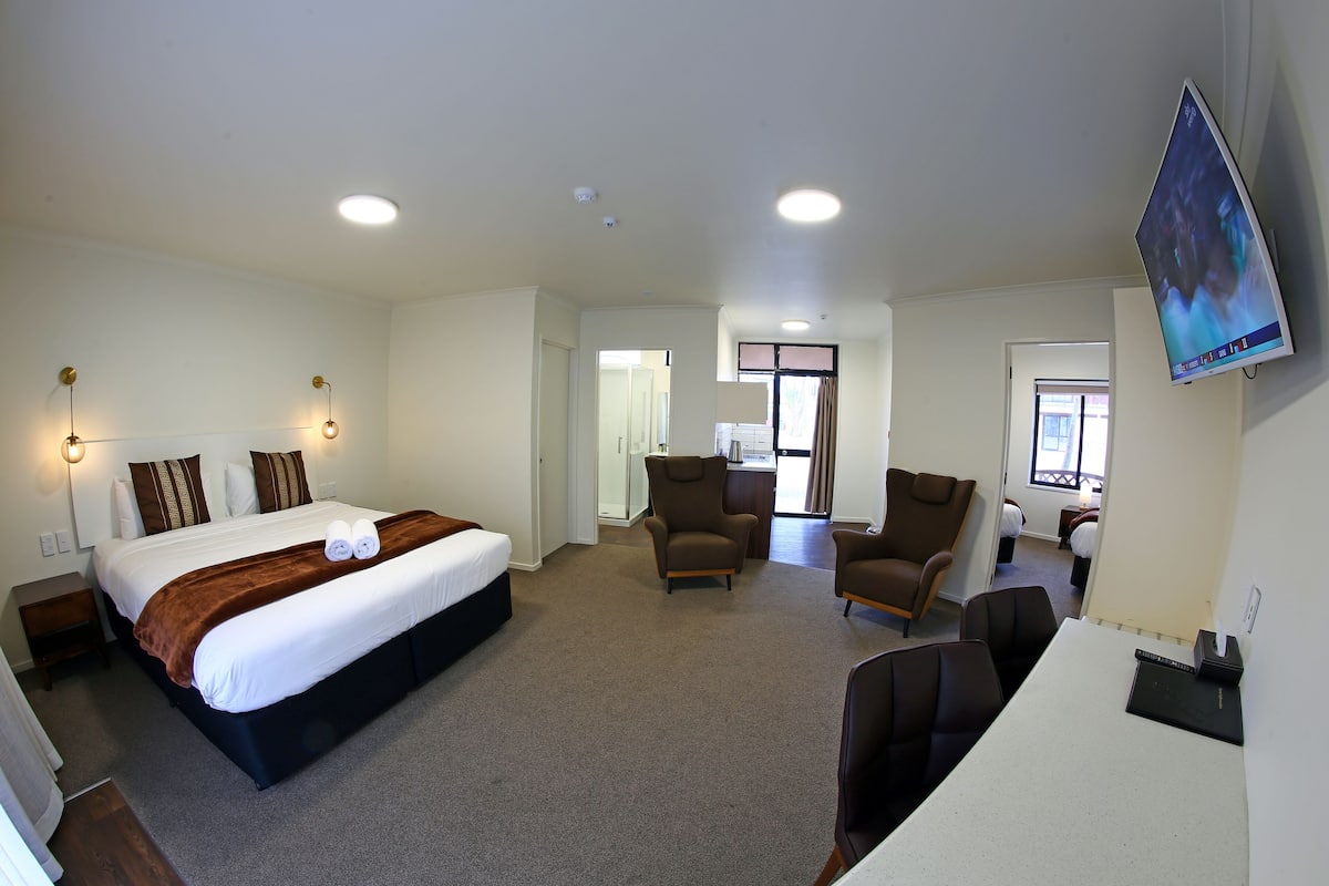 Ein Hotelzimmer mit einem großen Bett, zwei Sesseln, einem Schreibtisch und einem an der Wand befestigten Flachbildfernseher.