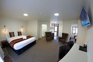 A hotel room with a large bed, two armchairs, a desk, and a flat-screen TV mounted on the wall. - Heritage Court Lodge  (Oamaru)