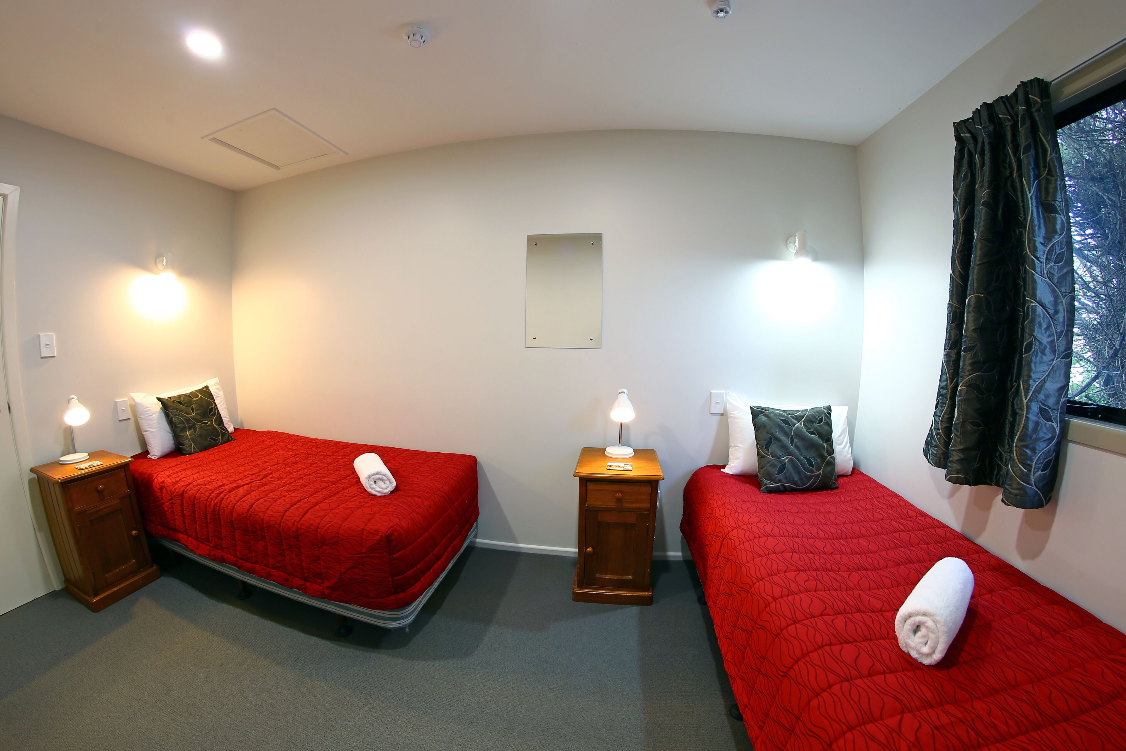 A hotel room with two beds, each with a red bedspread and pillows, a nightstand with a lamp, a small mirror on the wall, and a window with curtains.
