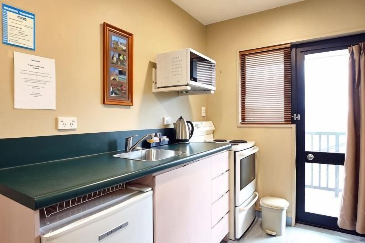 A kitchenette with a sink, stove, and microwave. There is a trash can and a door leading to a balcony.