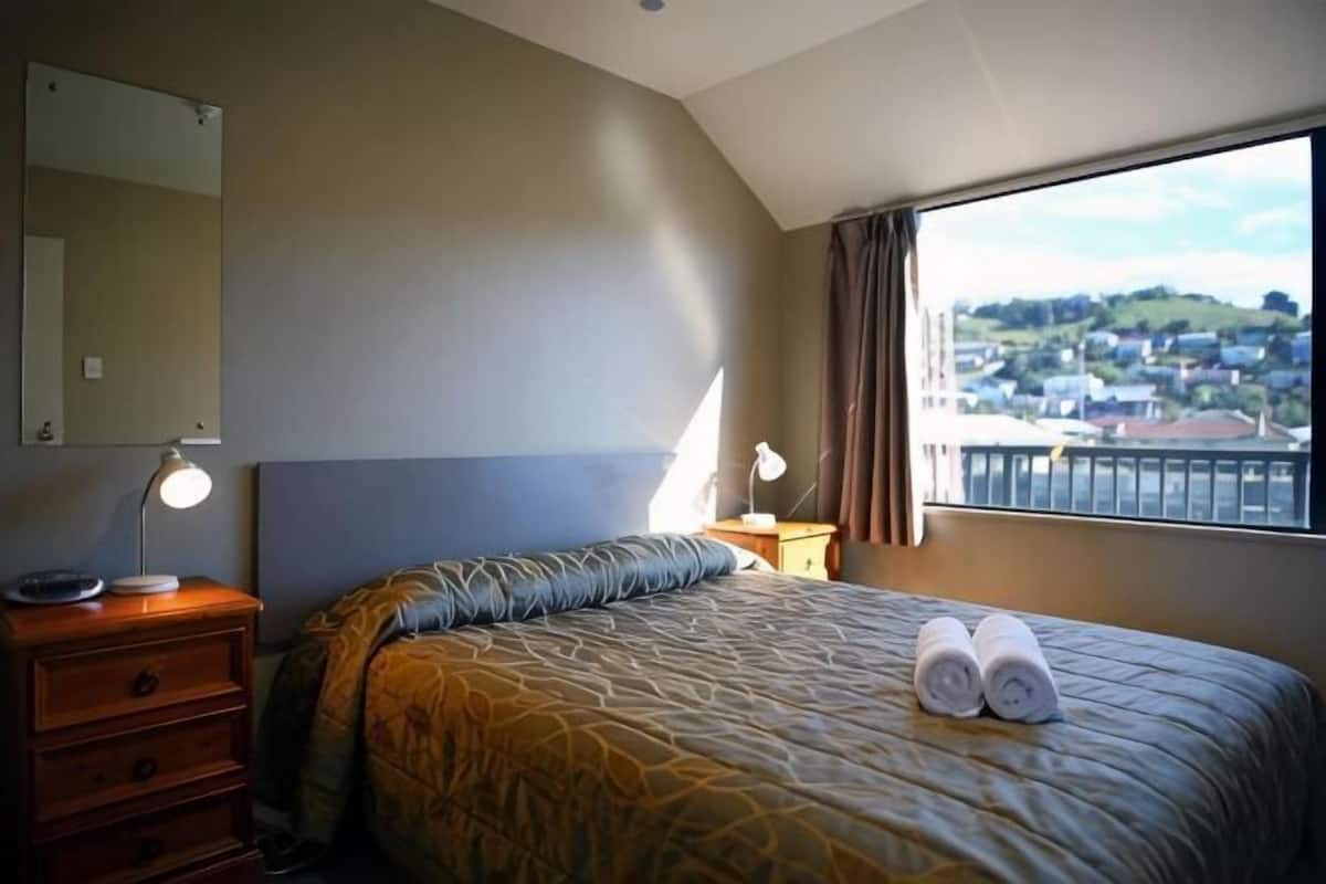 Ein Hotelzimmer mit Bett, Nachttisch, Lampe, Kommode und einem Fenster mit Blick auf eine Stadt.