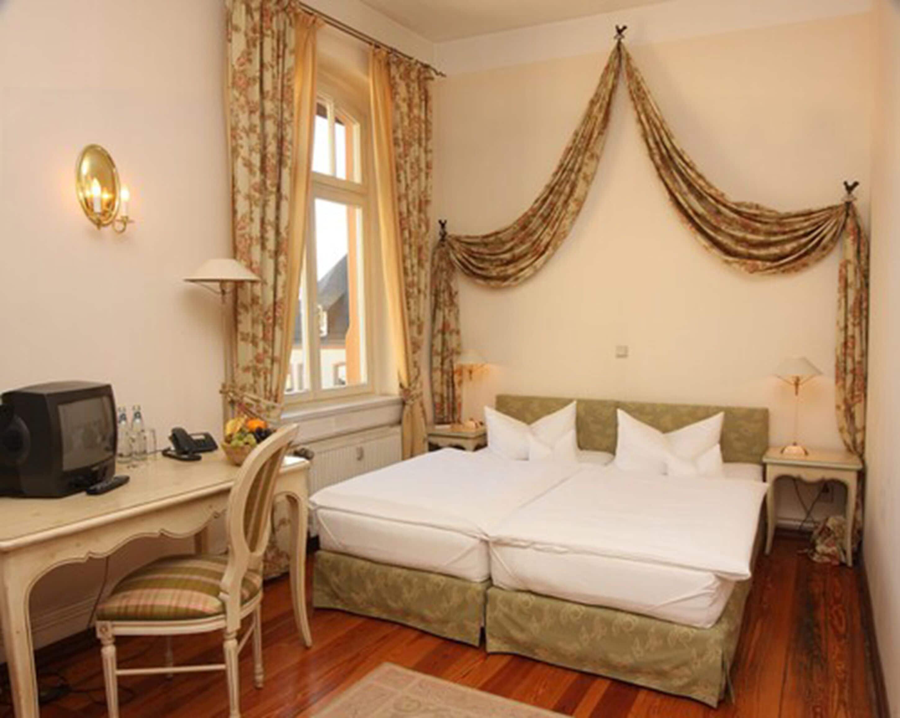 A hotel room with a double bed, a desk with a computer, a chair, and a window with curtains.