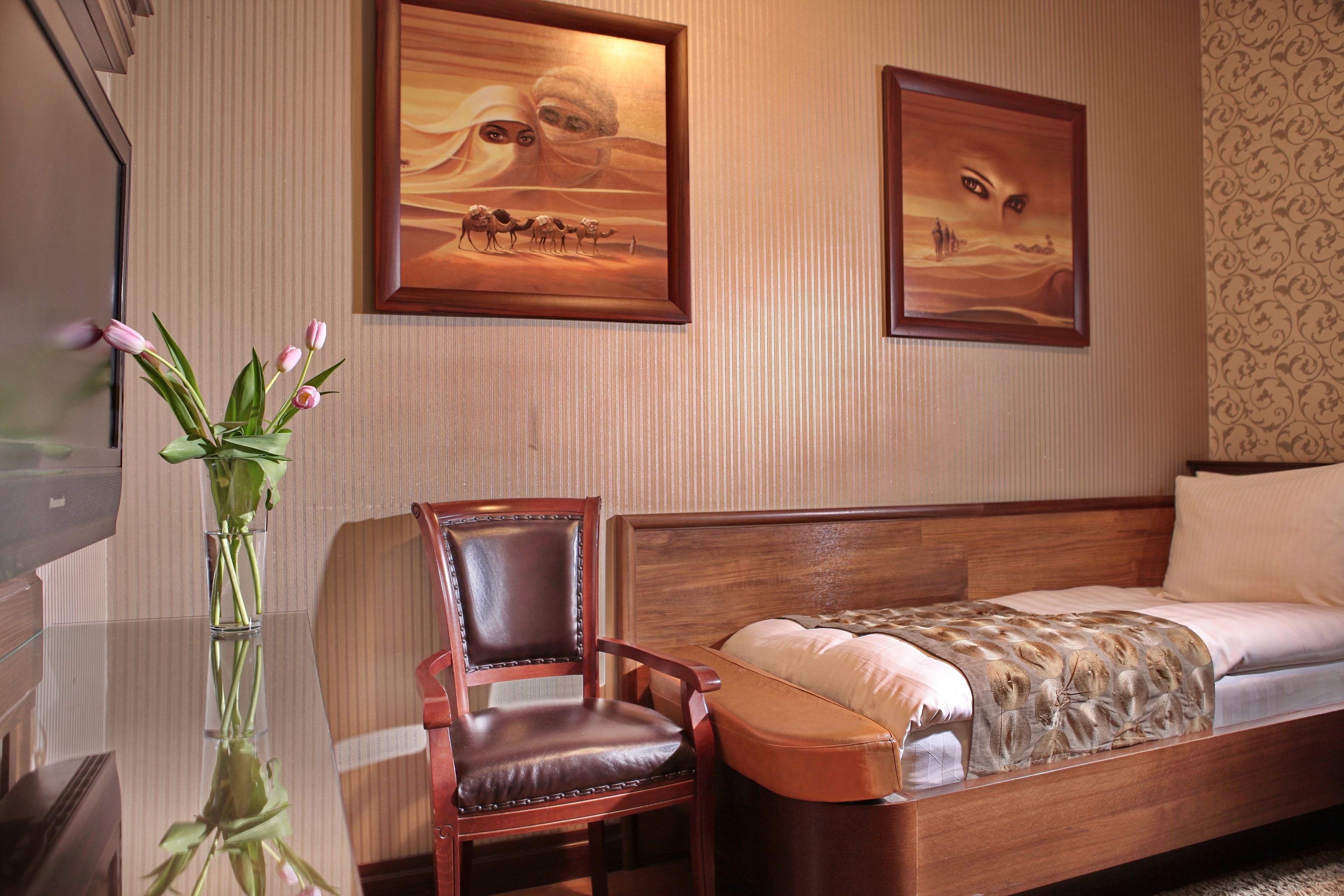 Une chambre d’hôtel avec un lit en bois, une chaise, une table en verre ornée d’un vase de fleurs et deux tableaux encadrés sur le thème du désert accrochés au mur.