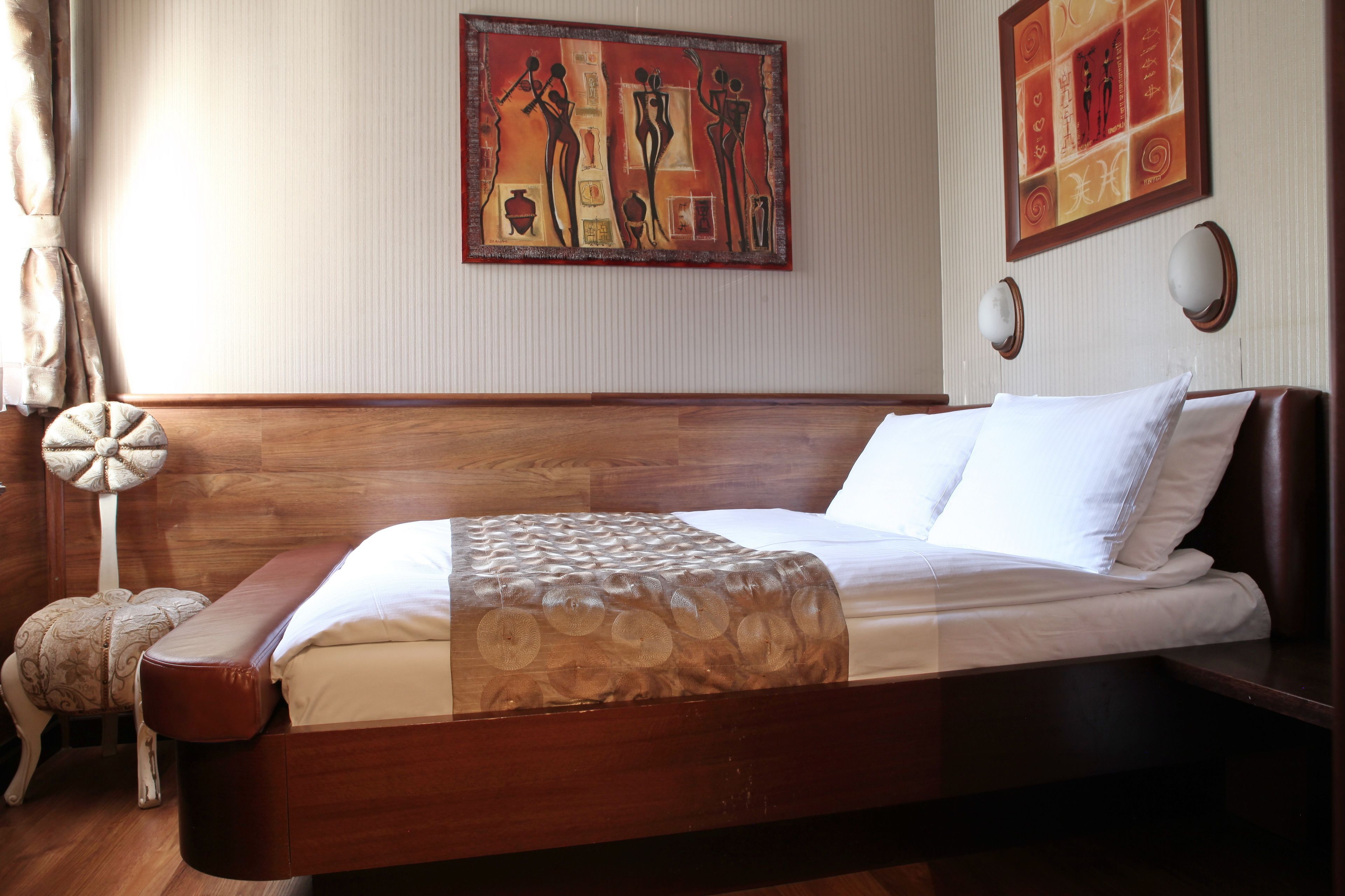 Una habitación de hotel con cama de madera, ropa de cama blanca, un cojín decorativo, una obra de arte colgada en la pared y una lámpara de noche.