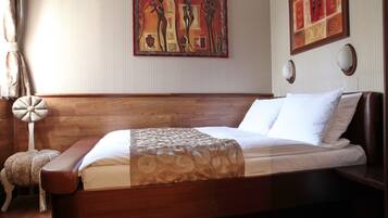 Una habitación de hotel con cama de madera, ropa de cama blanca, un cojín decorativo, una obra de arte colgada en la pared y una lámpara de noche.