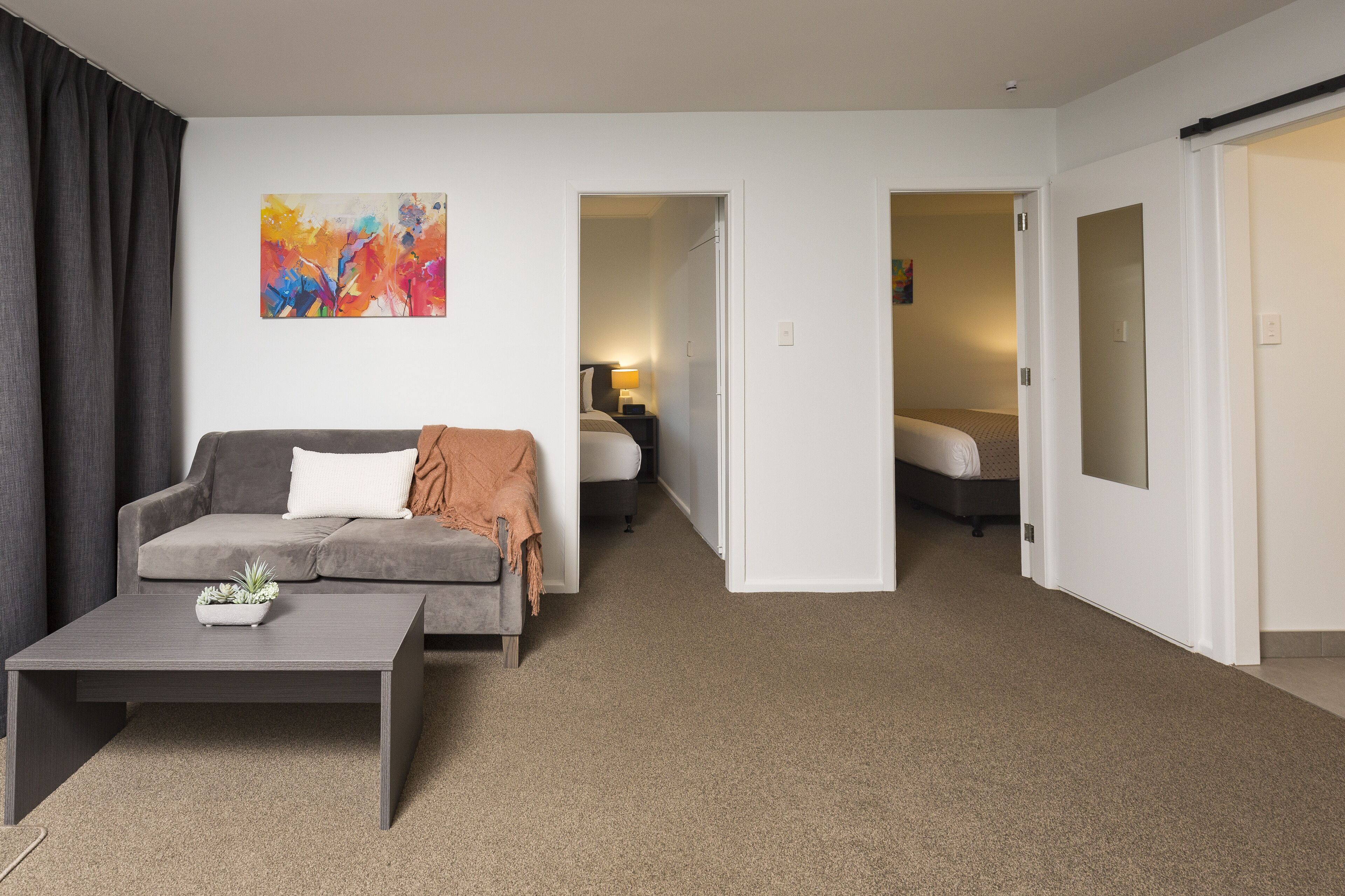 A modern hotel room with a grey sofa, a small coffee table with a plant, a colorful abstract painting on the wall, and a separate bedroom visible through an open door.