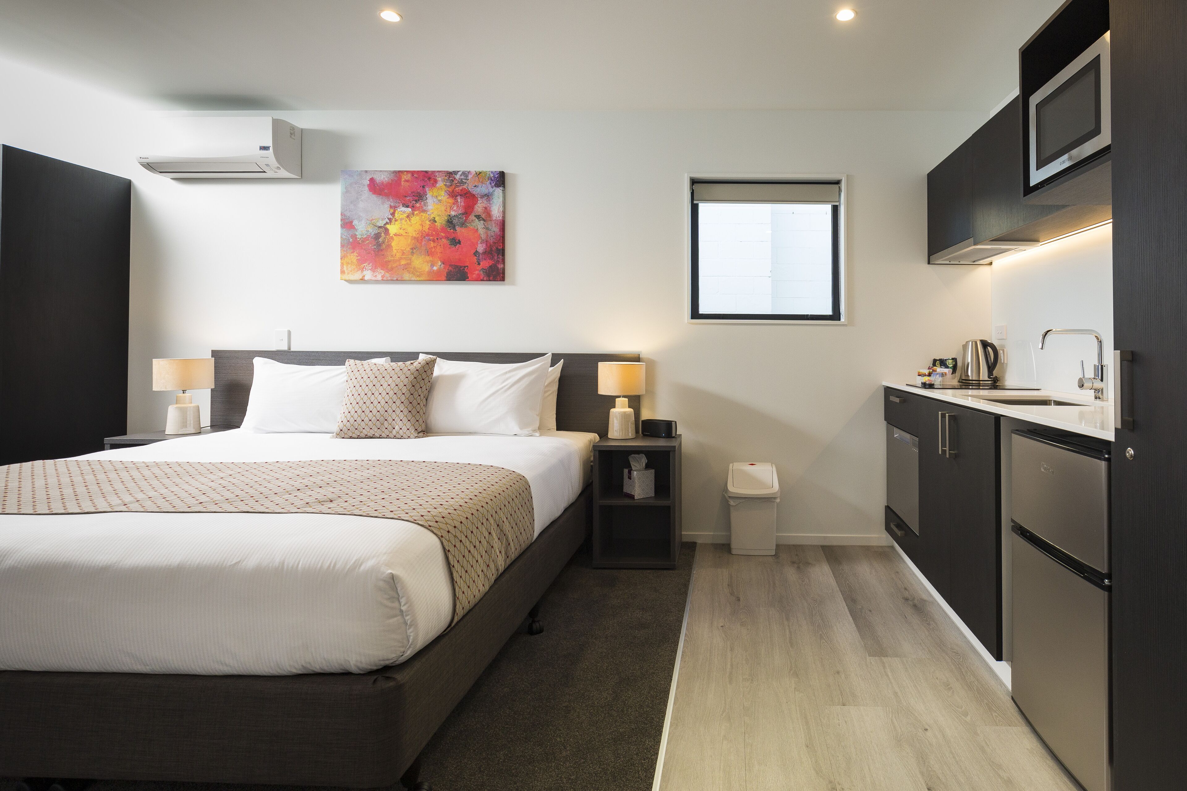 A hotel room with a bed, bedside lamps, a small table with a lamp, a trash bin, a microwave, and a window with blinds.
