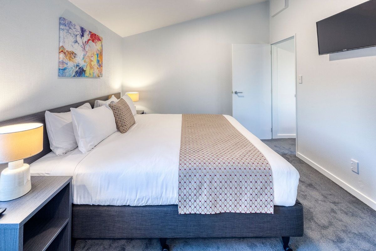 Een netjes opgemaakt bed met witte lakens en een decoratieve plaid, een bedlampje en een aan de muur bevestigde televisie.