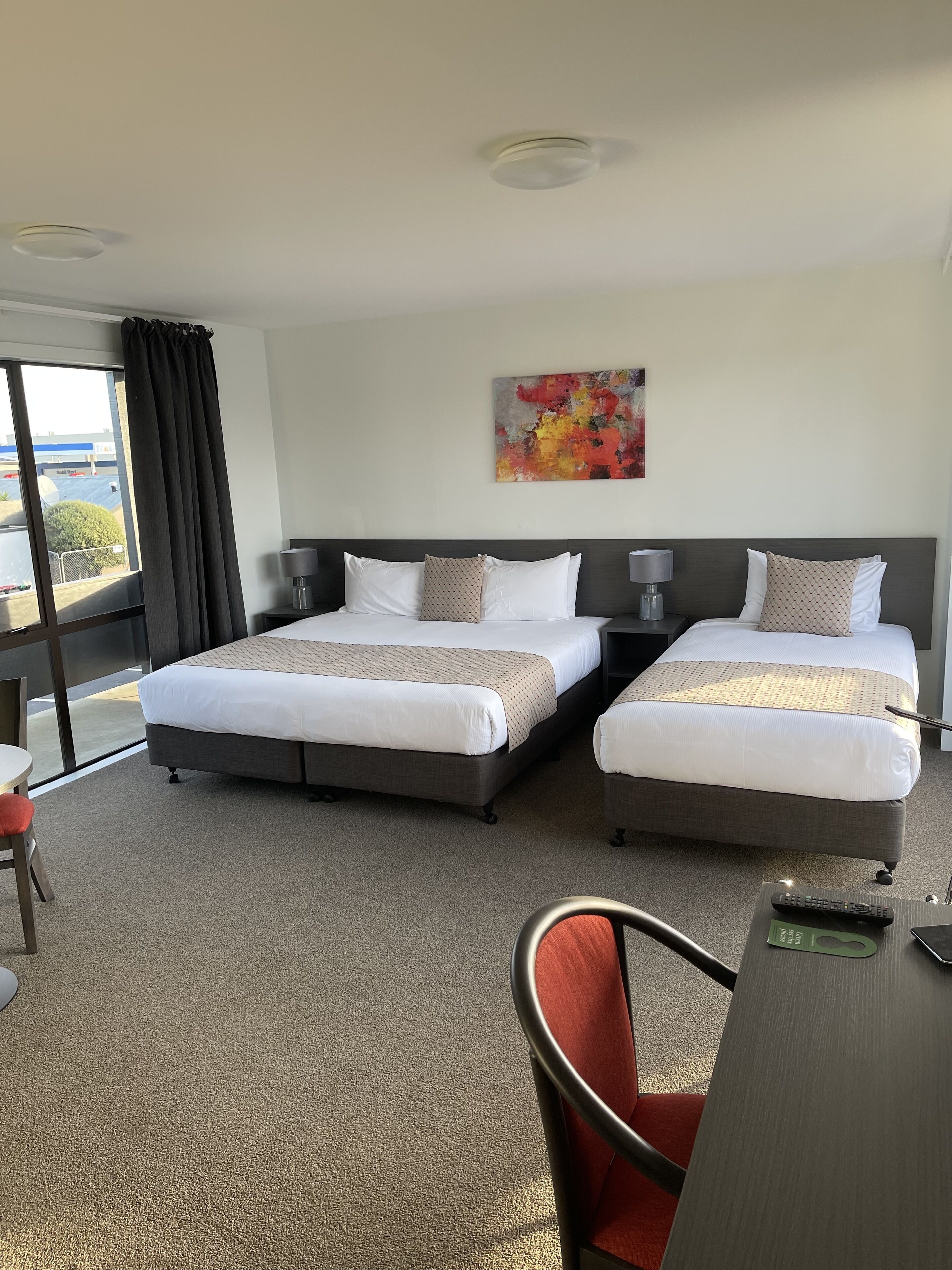 A hotel room with two beds, a large window with curtains, a painting on the wall, and a desk with a chair.