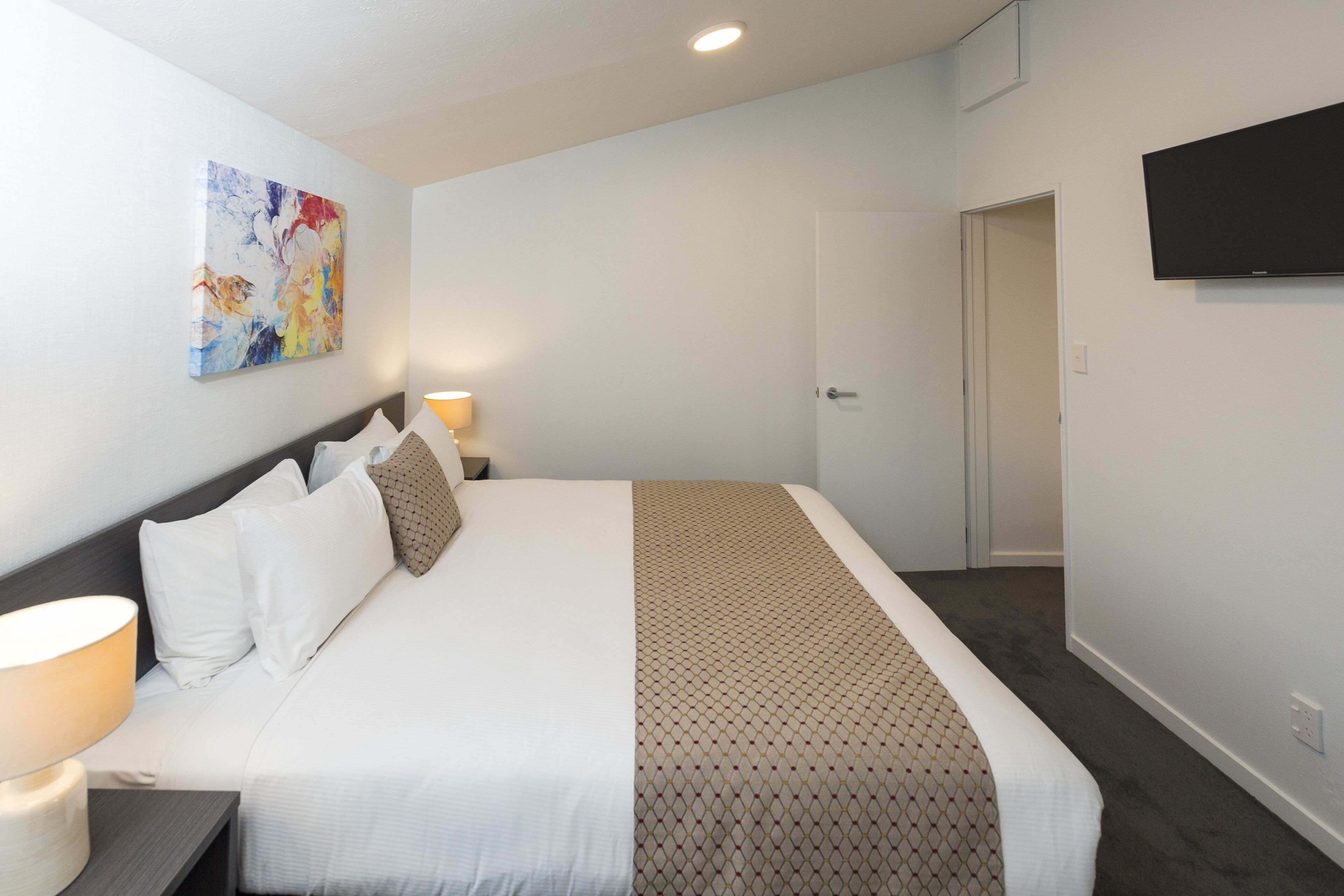Hotelkamer met een bed, een aan de muur bevestigde televisie, een deur naar een andere kamer en een schilderij.