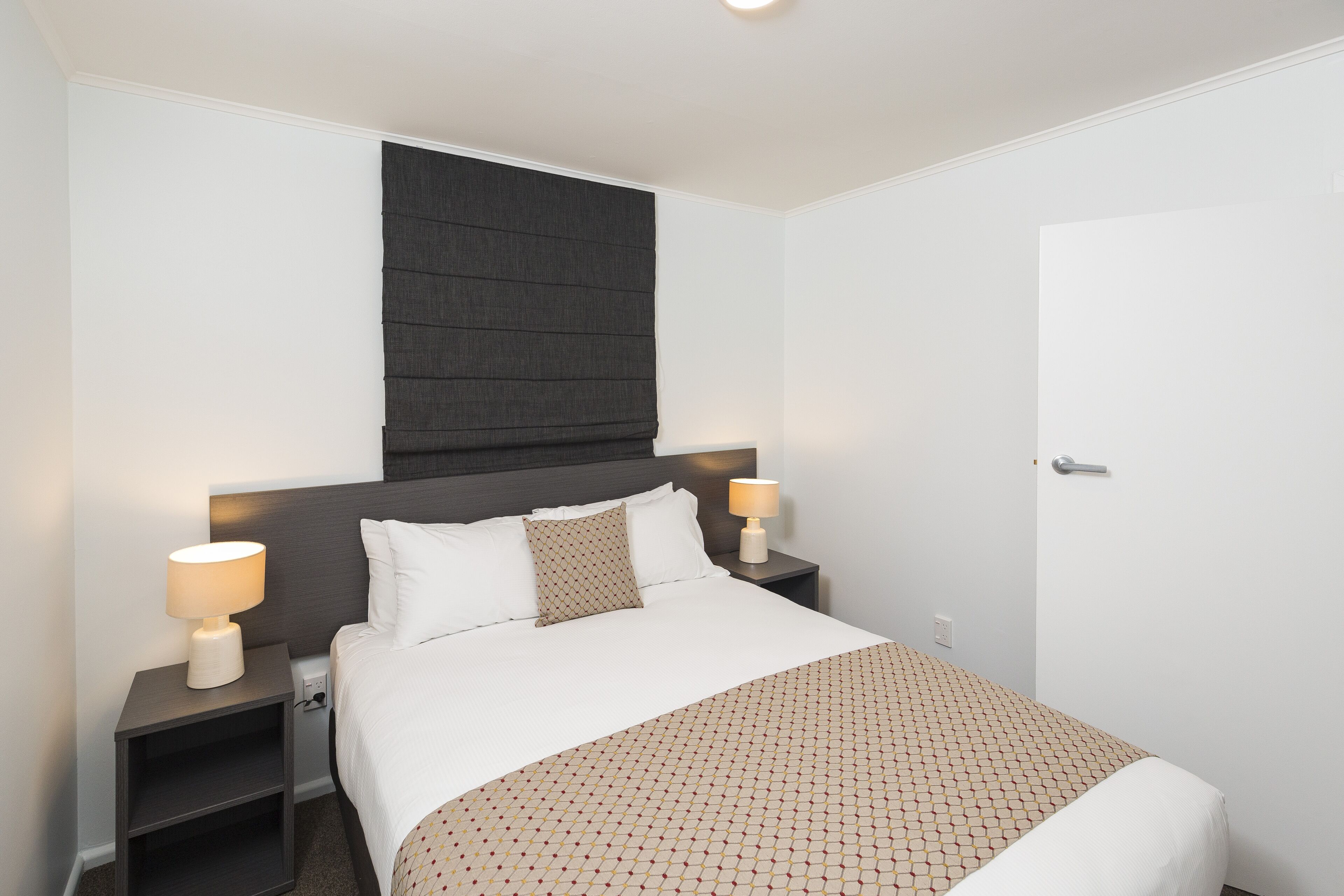 A hotel room with a bed, two bedside tables with lamps, a dark headboard, and a white door.