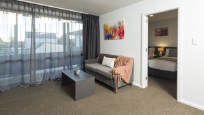 Habitació d'hotel moderna amb un sofà gris, una taula petita, una finestra gran amb cortines translúcides i un quadre a la paret.