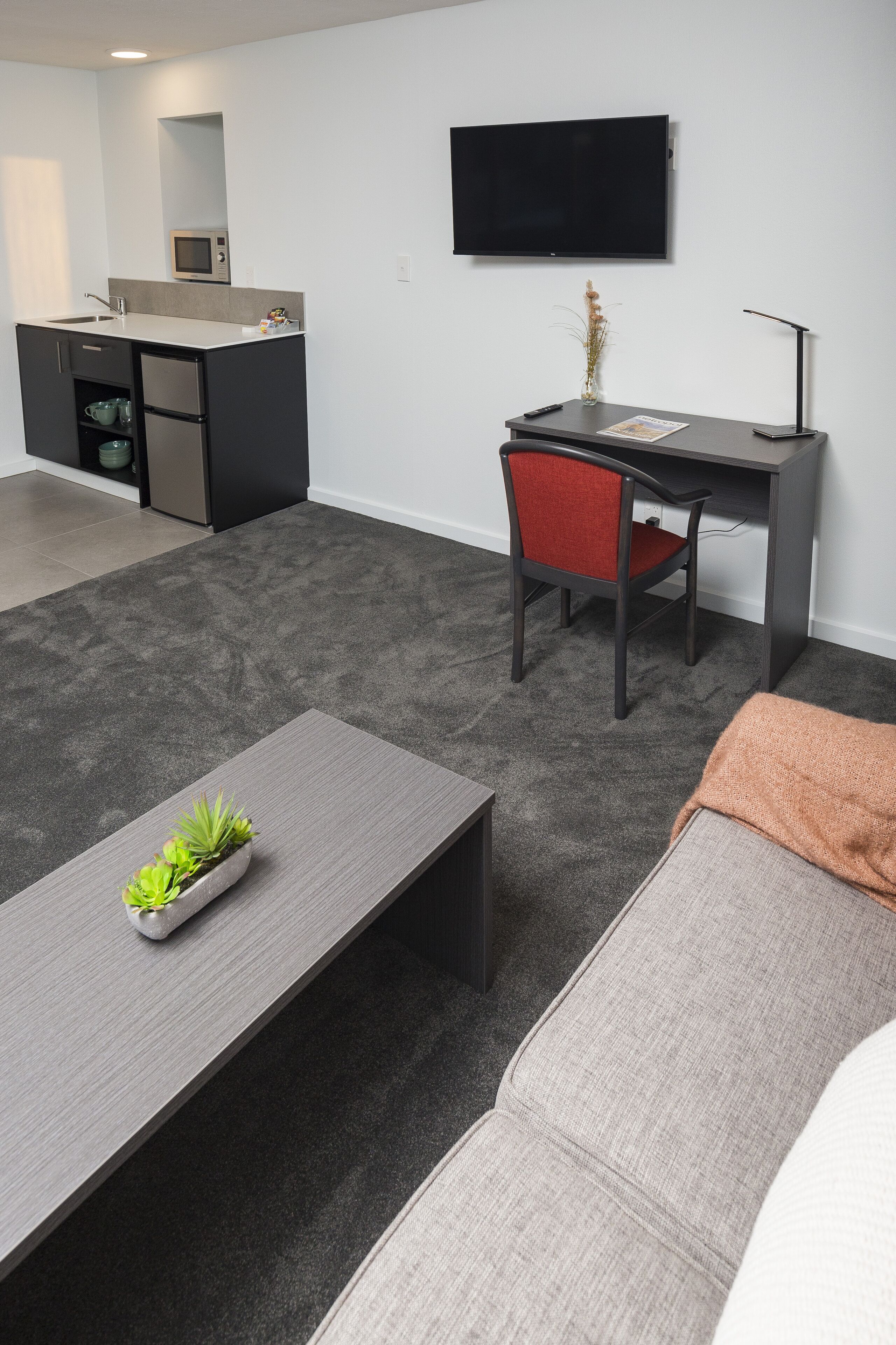 En moderne stue med en grå sofa, et lille bord med en plante, en rød stol, et skrivebord med tilhørende stol, et fladskærms-tv og et køkkenområde med en mikrobølgeovn.