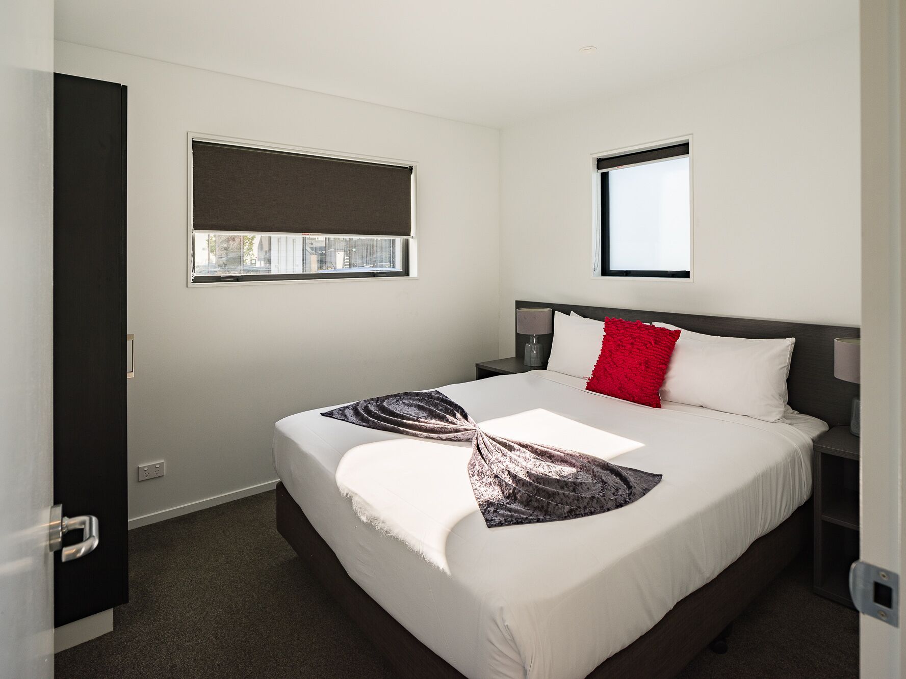 A modern hotel room with a large bed, a nightstand, a window with a blind, and a door.